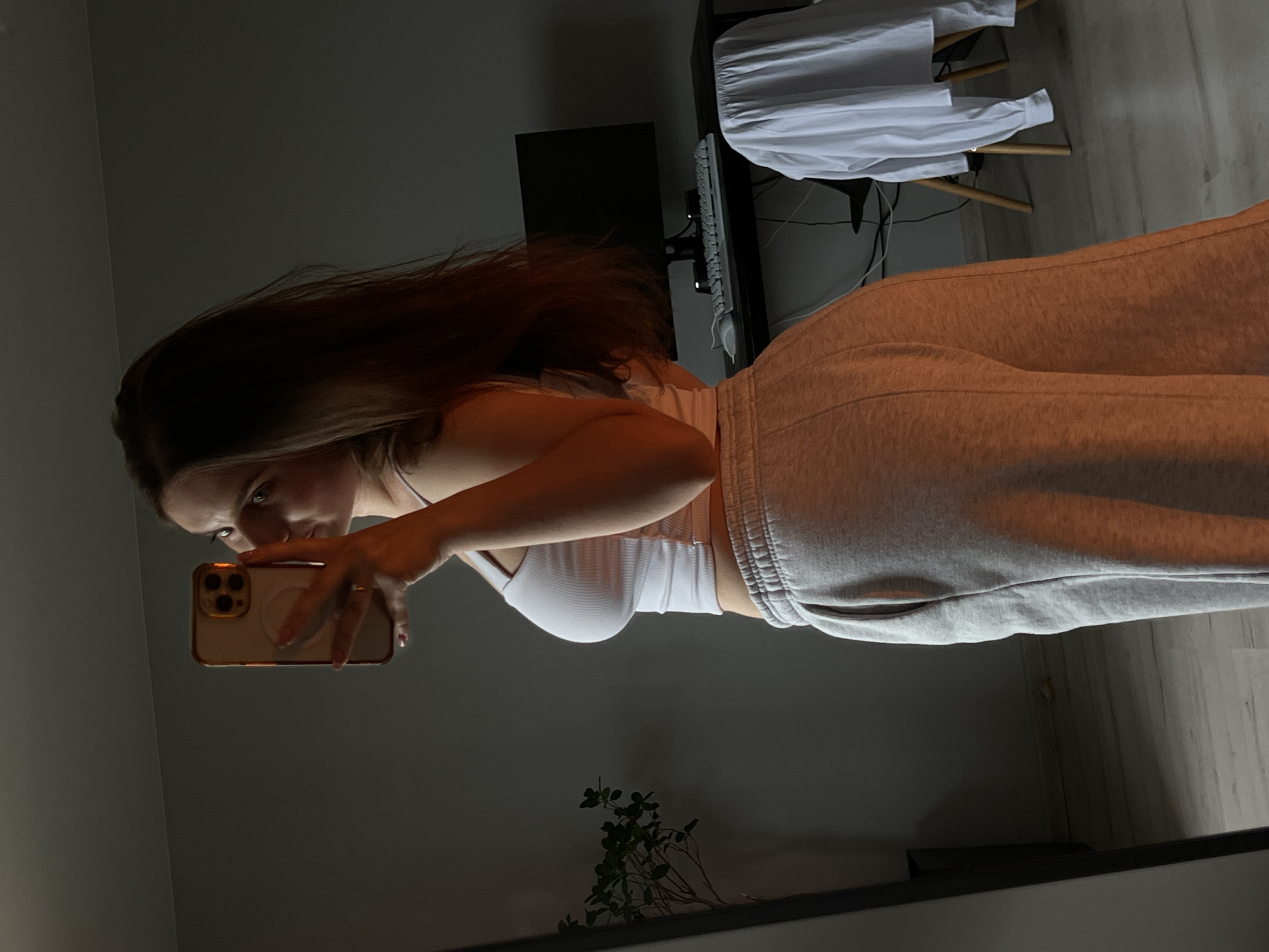 A female individual is depicted indoors, capturing a mirror selfie in an environment characterized by low, warm lighting. She has long, dark brown hair and is wearing a white ribbed crop top and light grey sweatpants. Her right arm is raised, holding a smartphone with a gold-toned case featuring multiple camera lenses and a circular grip accessory. A ring is visible on her right hand.
The background, visible both directly and in the mirror's reflection, includes a plain grey wall and light-colored wooden or laminate flooring. To the left, a small green plant in a dark pot is reflected. In the central and right portions of the background, a dark desk holds a black computer monitor, a white keyboard, and a white mouse. A chair with wooden legs, draped with a white long-sleeved shirt, is positioned beneath the desk. Dark cables are visible behind the desk.
There are no discernible visual elements within the image that indicate a specific geographical location such as Ternopil, Ukraine.
