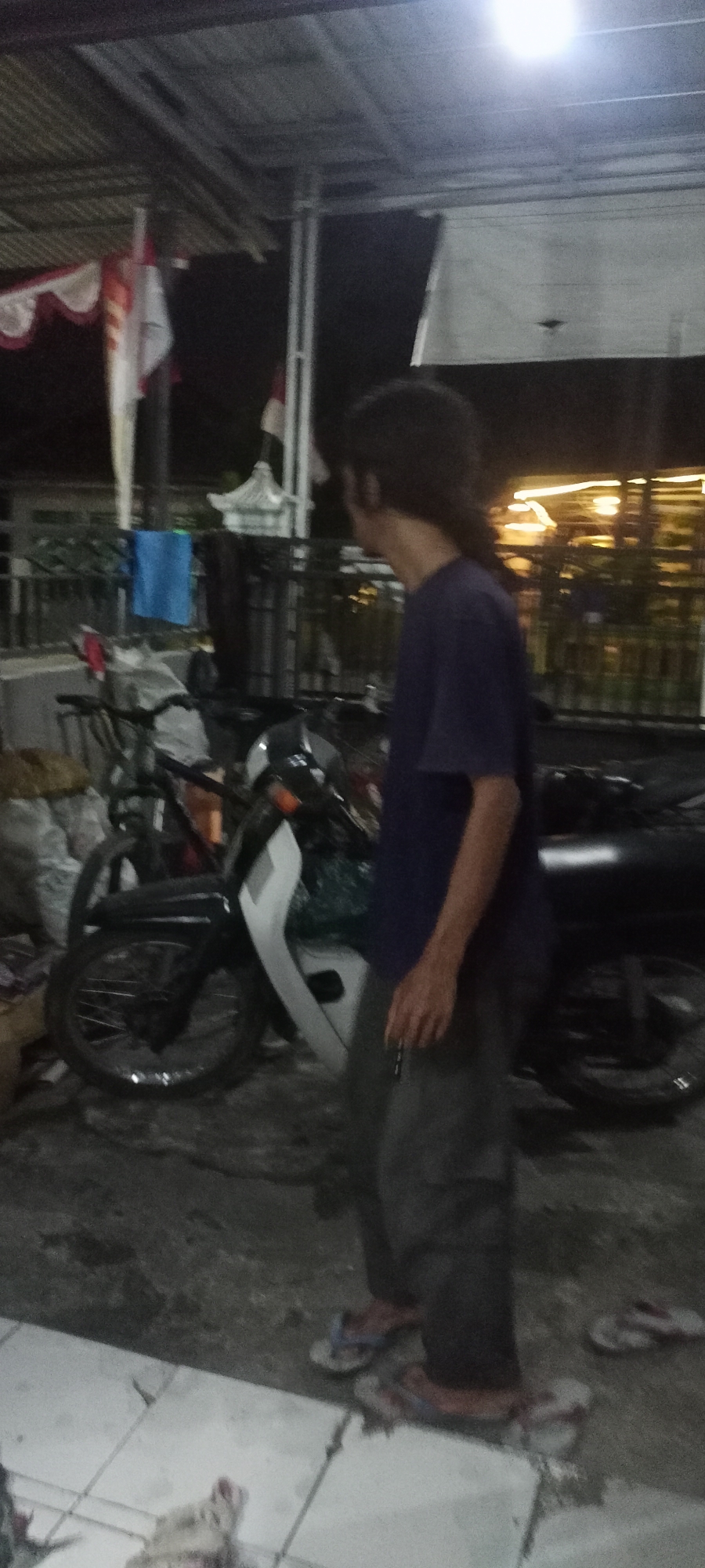 The image depicts an outdoor scene at night, likely in Gempolklutuk, Indonesia. A person with long, dark hair is standing and looking to the right. They are wearing a dark blue t-shirt and grey pants. The person is standing next to a motorcycle. In the background, there are flags, a building with a corrugated metal roof, and lights. There are also several bags and a blue cloth hanging in the background. The ground is paved with tiles in the foreground, transitioning to a rougher surface where the motorcycle is parked. The overall lighting suggests artificial light sources are present.