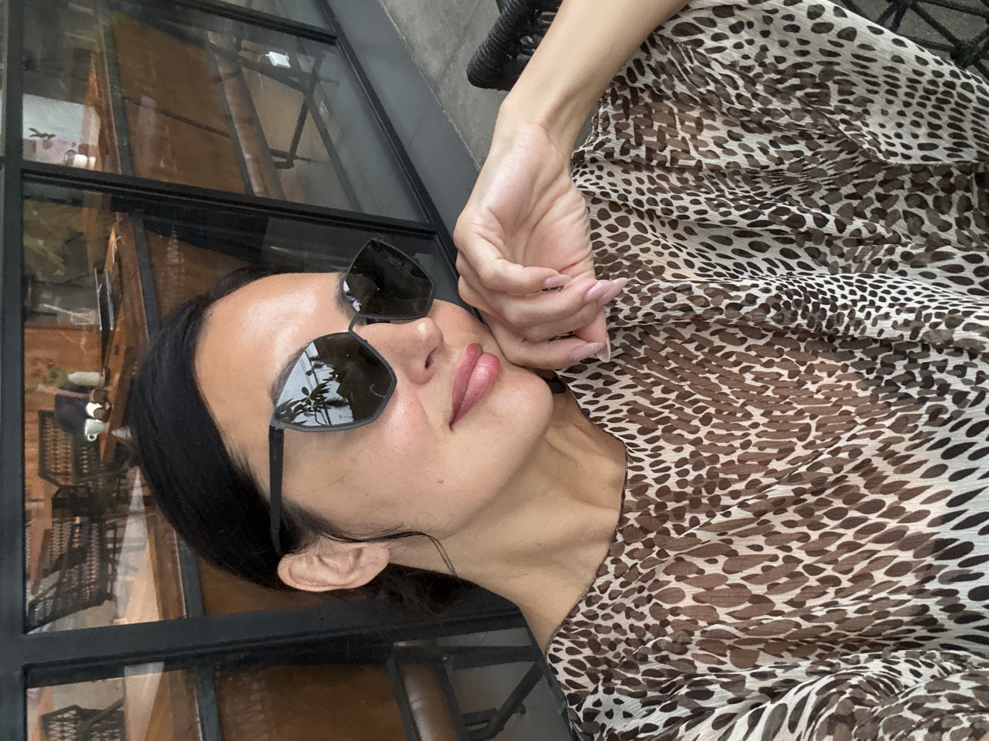 A woman, possibly of East Asian or Southeast Asian descent, is captured in a stylish, close-up portrait, likely in North Kuta, Indonesia. She is seen from the chest up, positioned slightly reclined or lounging next to a large window. Her dark hair is pulled back, revealing her ear. She wears black, reflective sunglasses that mirror greenery and bright surroundings. Her lips are subtly glossed with pink lipstick. A prominent feature is her outfit: a sheer, flowing top or dress with a distinct brown and white leopard or cheetah print. Her left hand, adorned with long, light pink manicured nails, rests delicately near her face. The background consists of black-framed window panes, reflecting indoor elements like wooden structures and hints of an outdoor view. The bright lighting indicates it is daytime. No text is visible in the image.