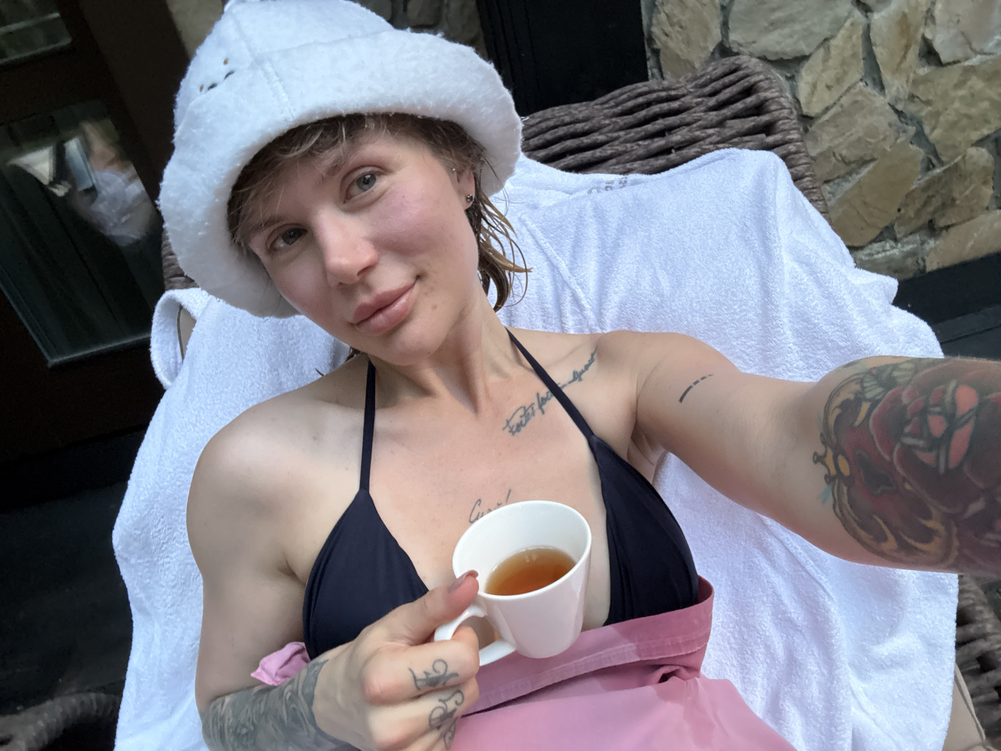 In Urlu-Aspak, Russia, a woman with fair skin and short, light brown hair is captured in a close-up, selfie-style photograph. She is wearing a white bucket hat and a black bikini top. Her left arm rests on a white towel, and her right hand is holding a small white teacup filled with amber liquid. The background shows a wicker chair and a stone wall. The woman has tattoos on her arms and chest, and a small silver earring is visible in her left ear. The image appears to be taken outdoors, possibly in a resort or spa setting.