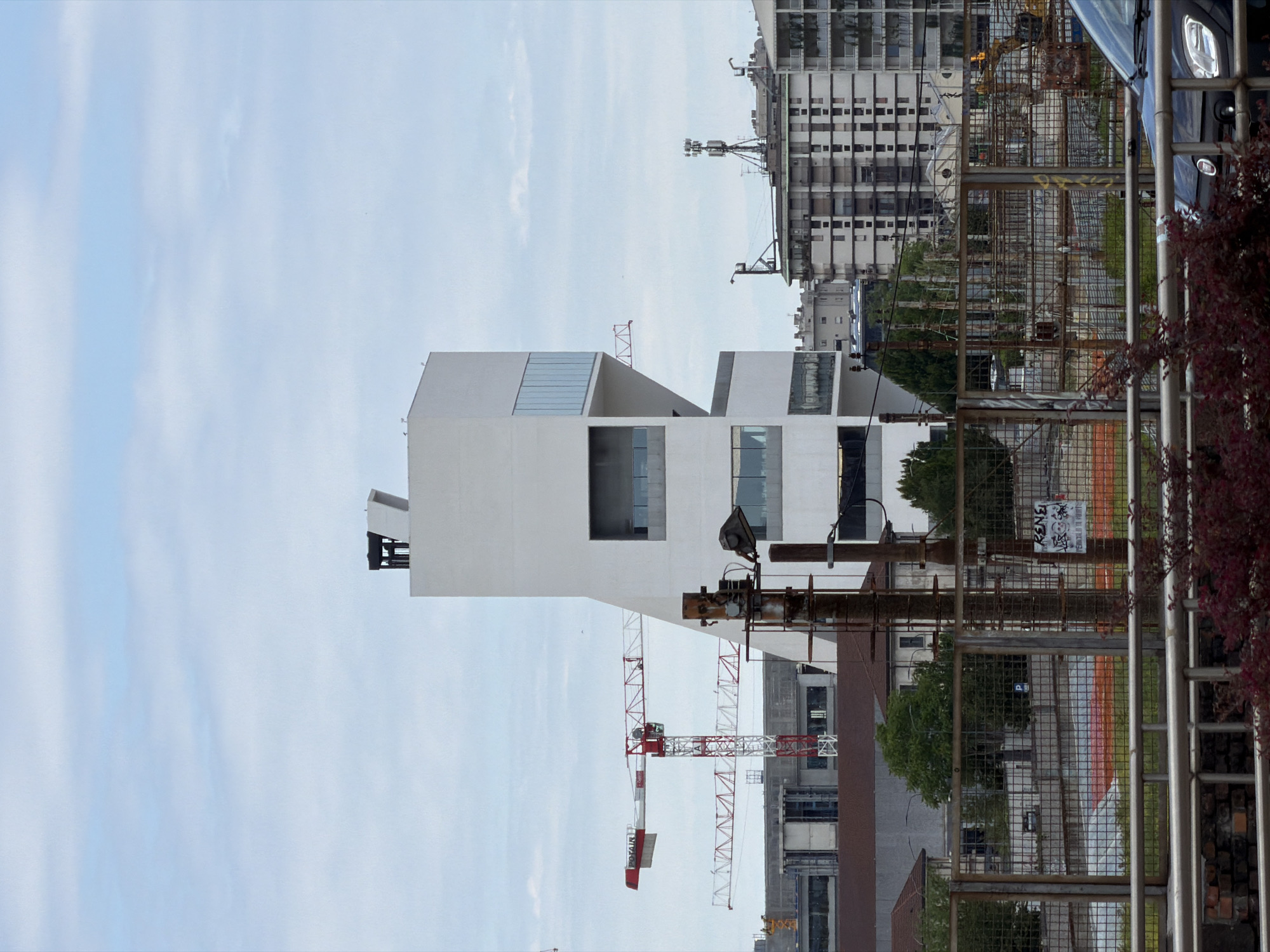 The image shows a view of Milan, Italy, featuring modern architecture, construction, and urban infrastructure.
A prominent white building with a geometric design and large windows is the focal point. A red and white construction crane stands nearby, indicating ongoing development.
In the foreground, a metal fence partially obscures the view. Behind the fence, there are trees, a road with orange stripes, and other buildings of varying heights and styles.
The sky is light blue with scattered clouds. A portion of a dark-colored car is visible on the right side of the image.