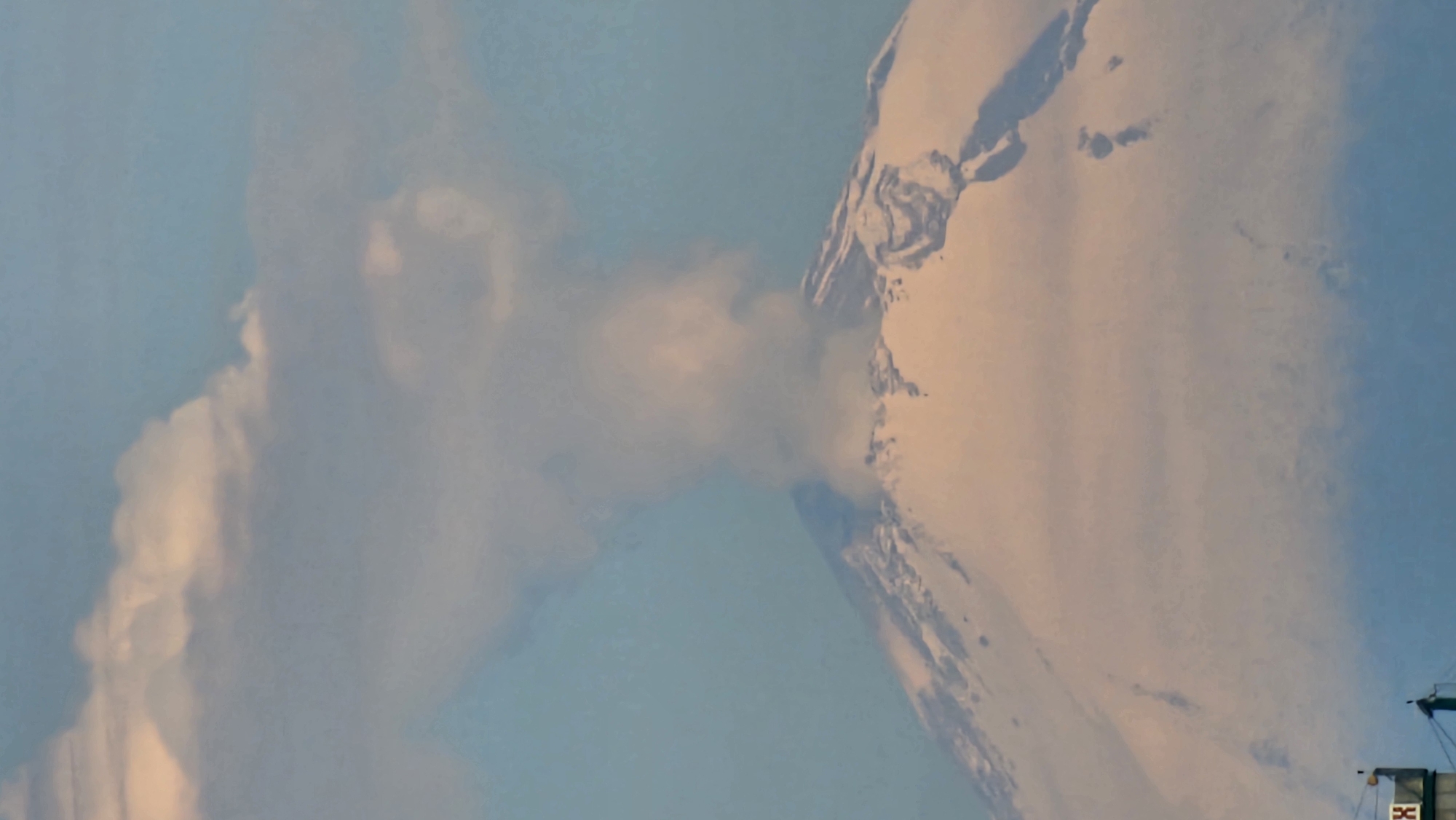 When correctly oriented, the image captures a dramatic volcanic event, likely involving Popocatépetl in Puebla, Mexico. A majestic, snow-capped mountain dominates the right side of the frame, its rugged, icy slopes illuminated by a warm, golden light, suggesting either sunrise or sunset.
A significant, light-colored plume of smoke, ash, or steam erupts vigorously from the mountain's flank, extending horizontally across the mid-ground and obscuring part of the peak. This dense plume merges with a larger, more diffuse cloud formation that occupies the left side of the sky, also catching the soft, warm light on its edges. The visible sky is a clear blue, providing a stark contrast to the dynamic volcanic activity.
In the bottom right corner, a small, dark-toned artificial structure or piece of equipment is partially visible, featuring rectangular elements. Faint white lettering, including a clear "X" mark, can be discerned on this structure. The scene powerfully conveys the raw force of nature, with the golden hour lighting adding a dramatic and almost ethereal quality to the erupting volcano.