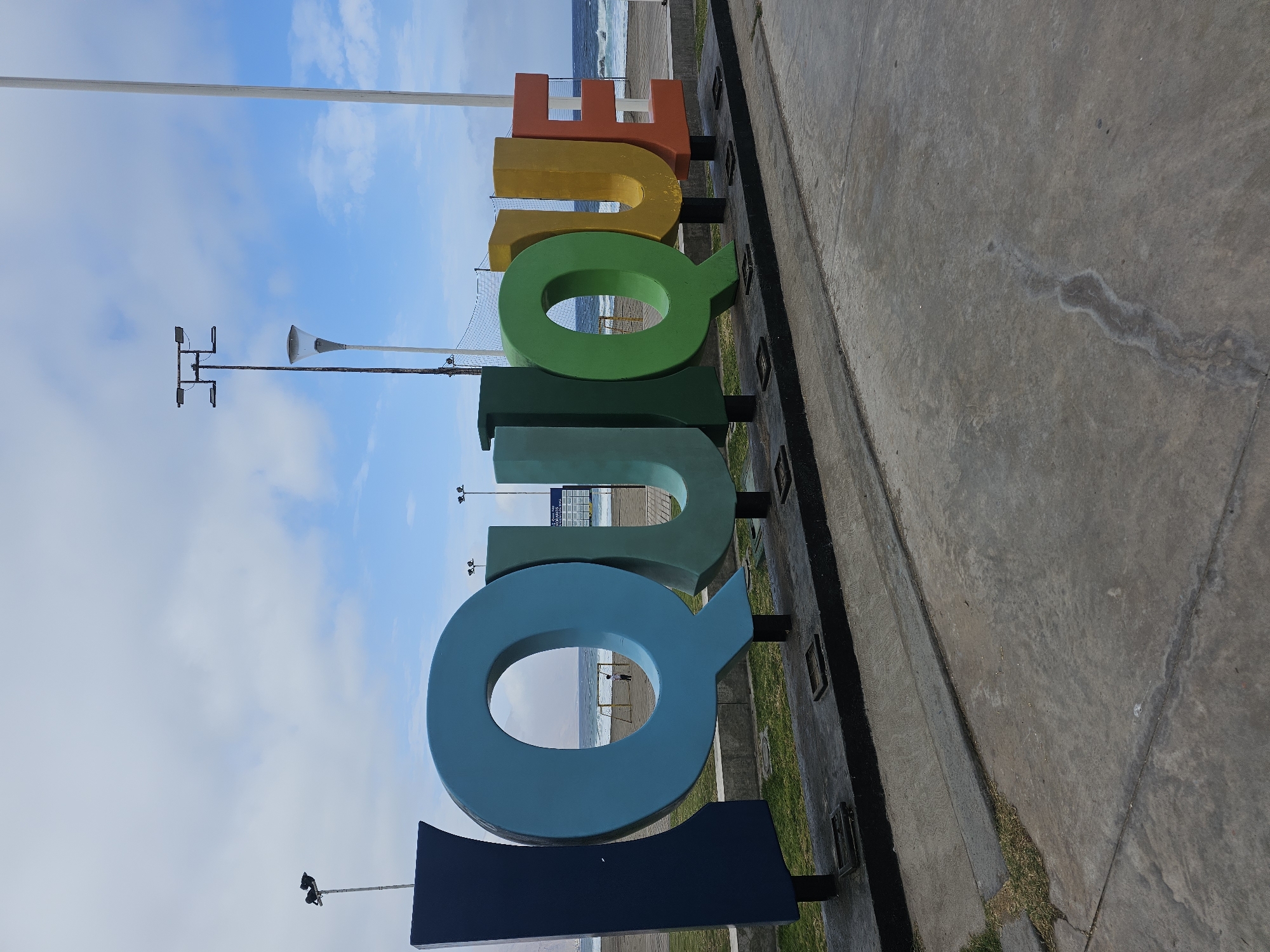 The image depicts a large, colorful sign spelling out "IQUIQUE" in front of the ocean. The letters are a deep blue, light blue, green, yellow, and orange, set against a backdrop of a cloudy sky. The sign is positioned on a concrete platform, with grass and a sidewalk leading up to it. In the background, the ocean waves crash onto the shore, and a few beachgoers can be seen in the distance. The overall scene suggests a coastal city known for its beaches and vibrant atmosphere.