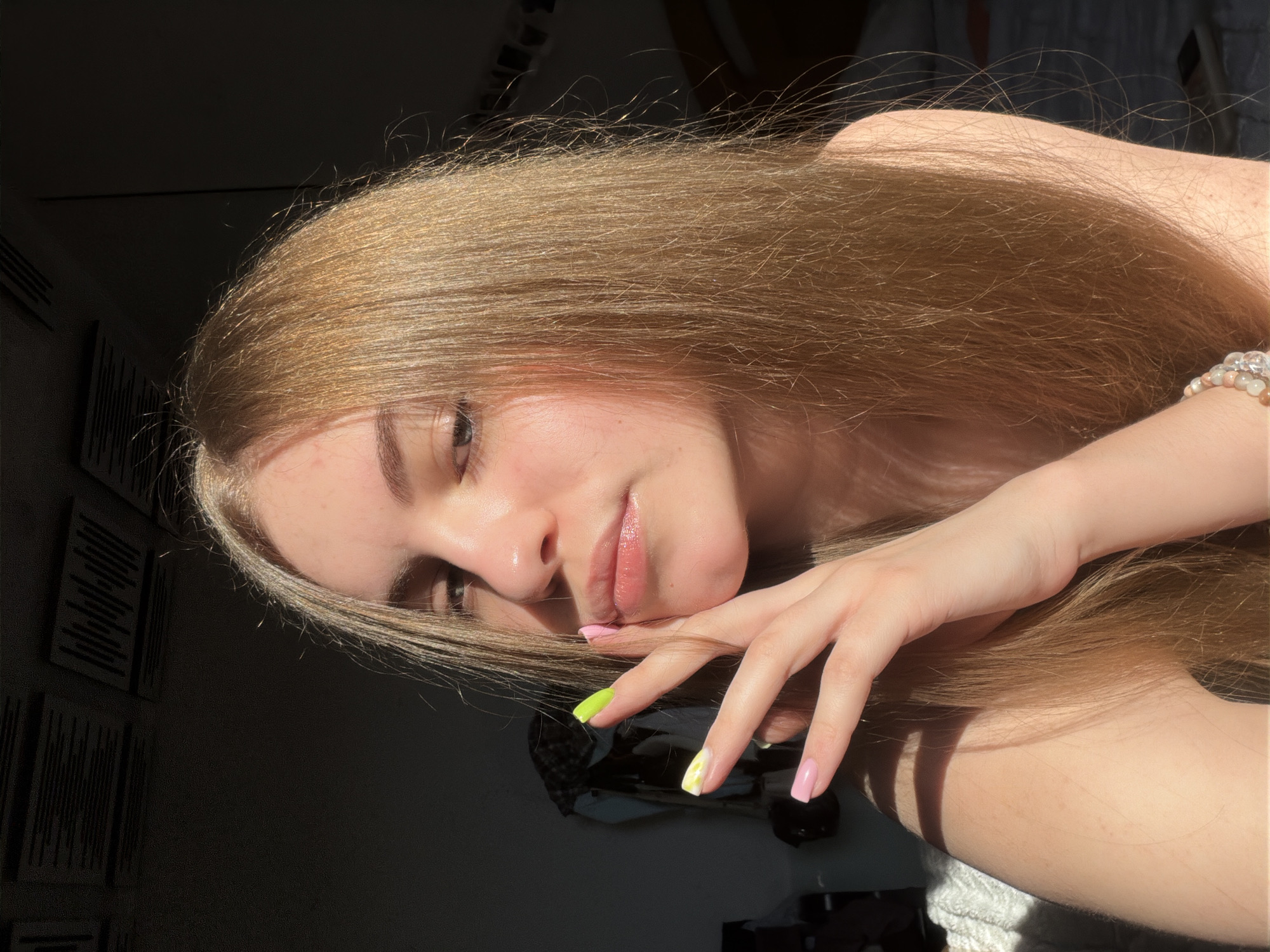 A young woman with long, light brown hair is captured in a close-up shot, her face turned towards the camera. Sunlight casts dramatic shadows across her features, highlighting her eyes and glossy lips. Her right hand, adorned with colorful nail polish, is raised to her face, with fingers delicately touching her cheek. She appears to be indoors, in a room with dark walls and what looks like acoustic panels on the ceiling. A beaded bracelet is visible on her left wrist. The setting appears to be a private residence. There is no indication that this image was taken in Dnipro, Ukraine.
