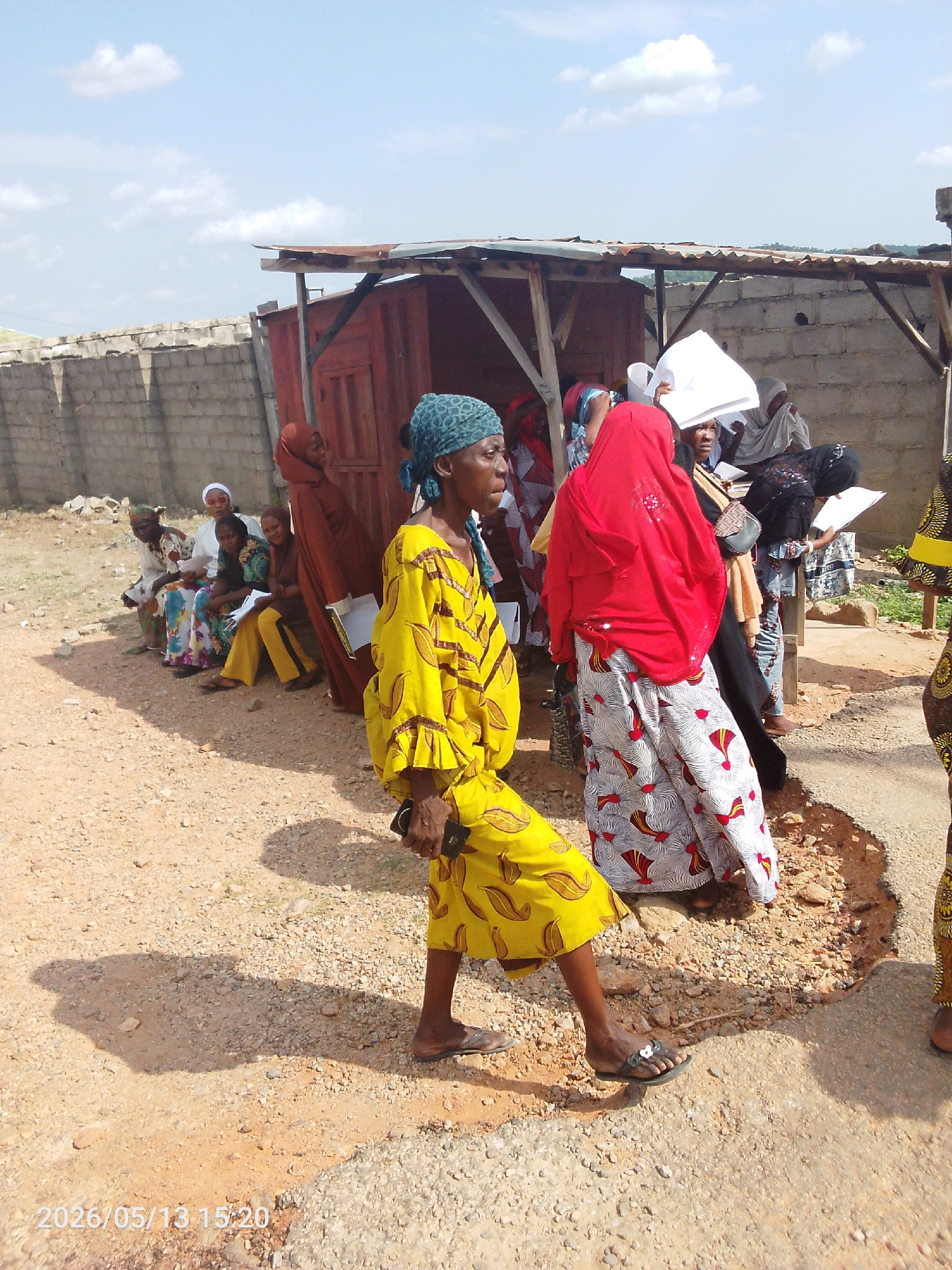 In Lokoja, Nigeria, a group of women are gathered outdoors on a partly cloudy day. One woman, wearing a yellow patterned dress and blue head covering, walks in the foreground, holding a cell phone. Nearby, another woman is wearing a red headscarf and a long, patterned skirt. Several other women are either standing or sitting by a makeshift structure with a corrugated metal roof and wooden supports. Some of the women are holding papers, possibly forms or documents. The ground is a mix of dirt and gravel. A concrete block wall is visible in the background. The timestamp "2026/05/13 15:20" is displayed in the lower left corner of the image.