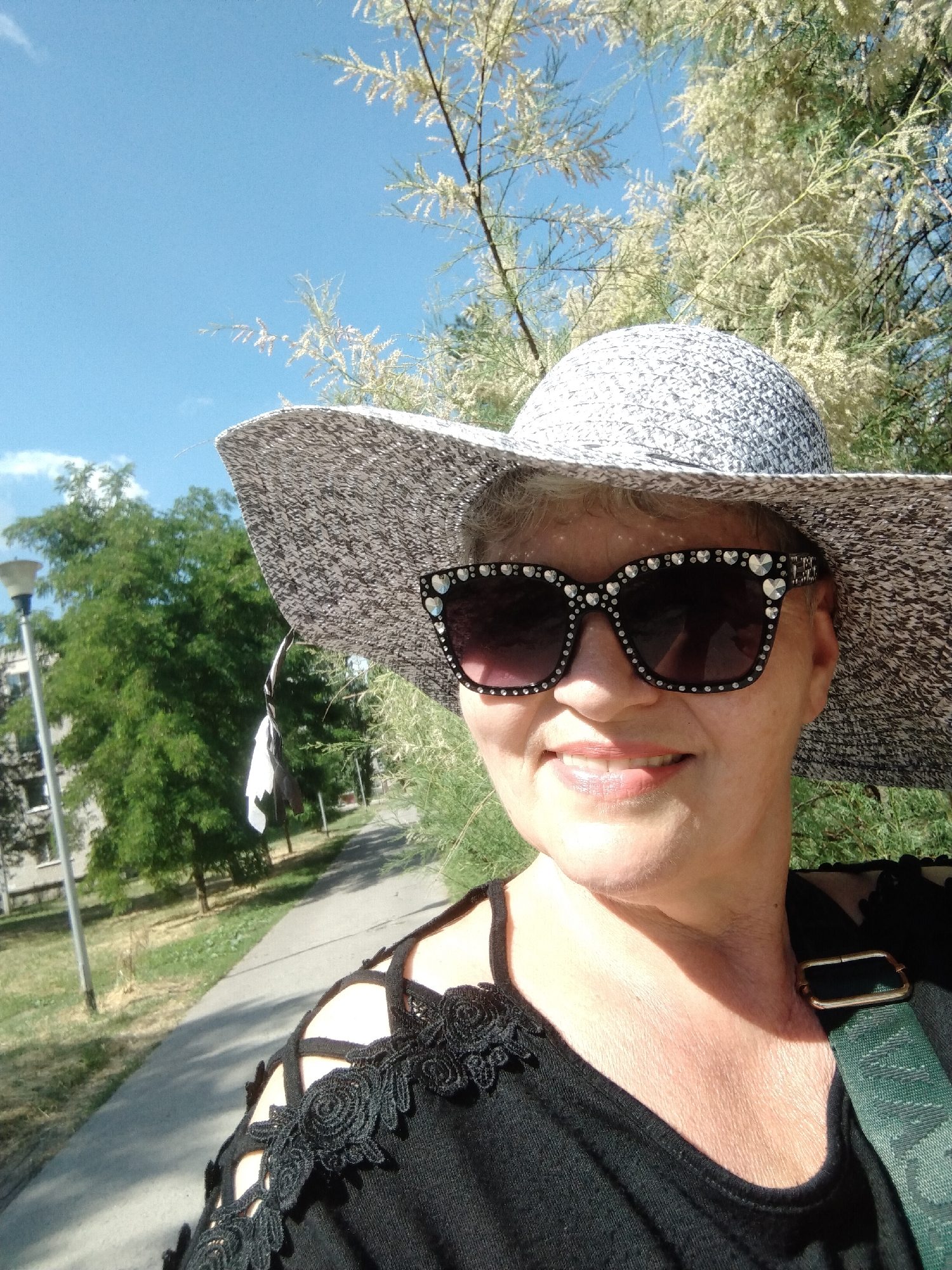 A woman is taking a selfie outdoors in Horishni Plavni, Ukraine. She is wearing a wide-brimmed patterned hat and large, embellished sunglasses. She has on a black top with lace accents on the shoulders. A green strap with a gold buckle is visible across her shoulder. Behind her, a paved walkway borders a grassy area with trees and a lamppost. The sky is blue with some clouds, and there are tree branches with small white flowers in the foreground.