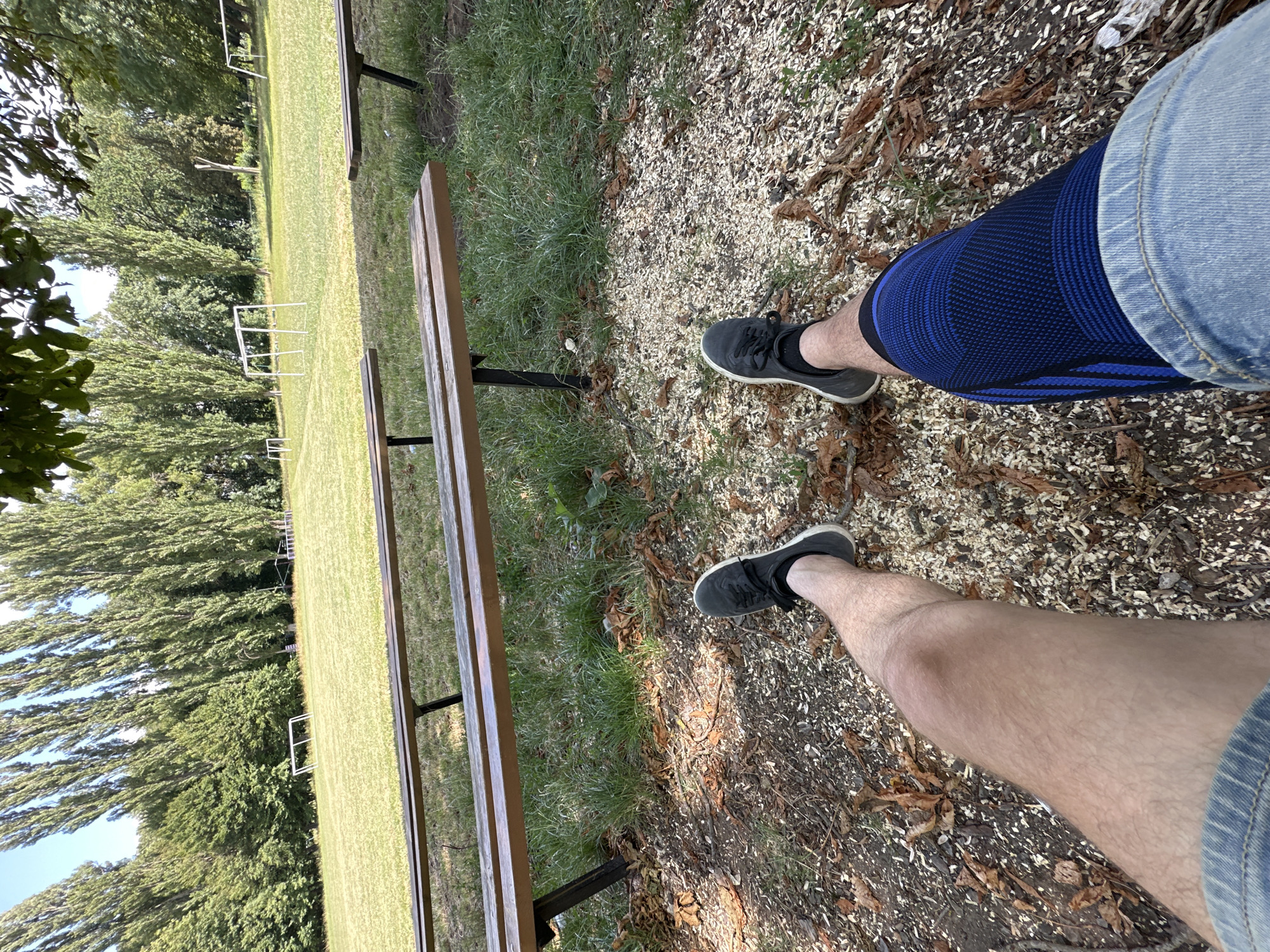 The image shows a first-person perspective of a person sitting on a wooden bench in an outdoor recreational area, possibly a park or sports field, in the city of Makiv, Ukraine. The person's legs are visible, wearing denim shorts, black athletic shoes, and a blue knee brace on the right leg. The ground is covered with wood chips and fallen leaves. In the background, a well-maintained green lawn is visible, with several small soccer goals scattered across it. Tall trees line the perimeter of the field, and a clear blue sky is above. The overall scene suggests a relaxed atmosphere on a sunny day.
