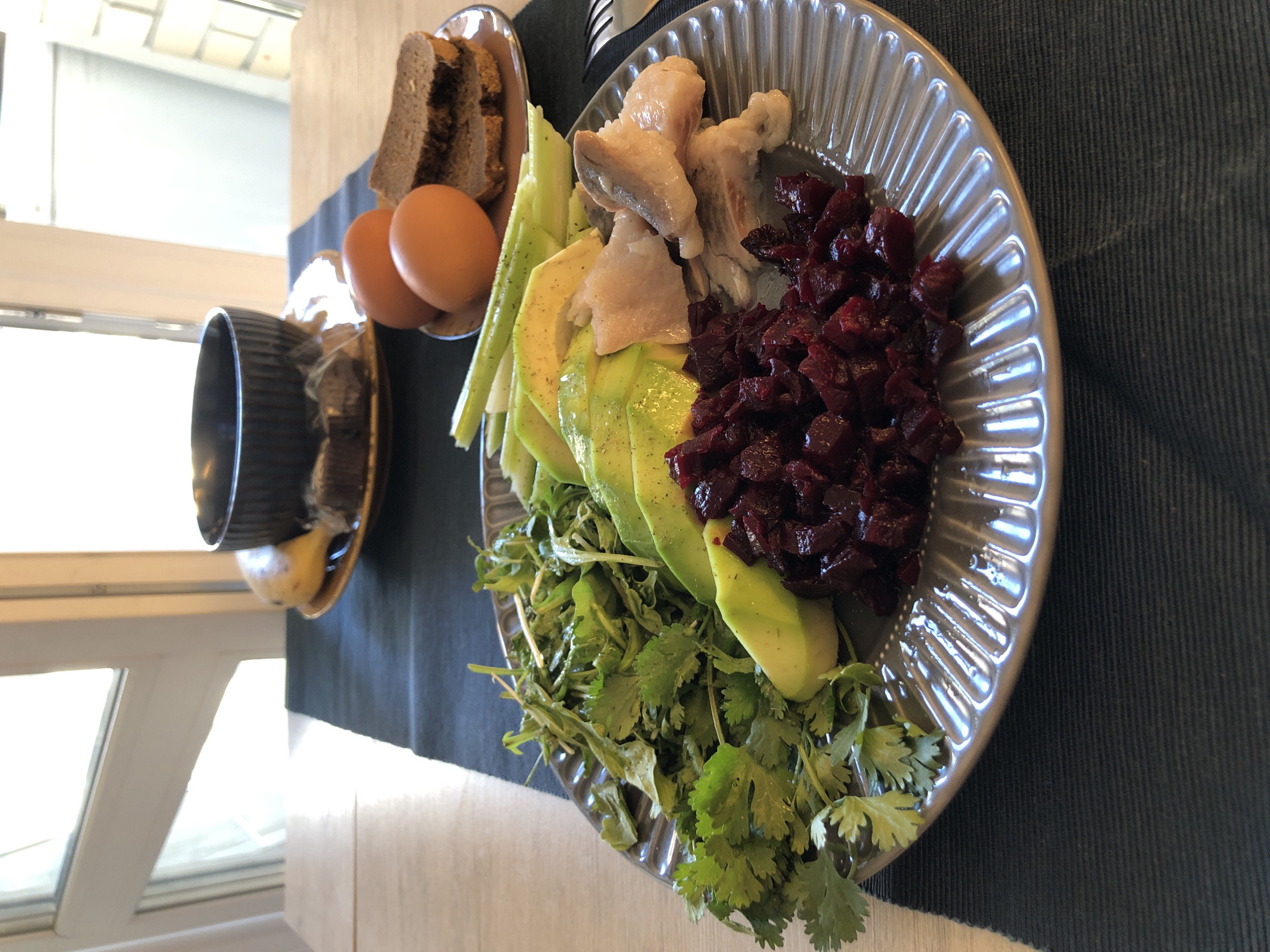 The scene is an interior dining setting in Saint Petersburg, Russia, featuring a prepared meal laid out on a light-colored wooden table. A dark gray, horizontally ribbed placemat or runner is centrally positioned on the table.
In the foreground, a large gray ceramic plate with a radiating ribbed pattern holds a selection of food items. On the left side of the plate, a pile of fresh green herbs, comprising both cilantro and arugula, is present. Adjacent to the herbs are several slices of avocado, followed by multiple diagonally cut celery stalks. Small dark specks are visible on the avocado and celery. Towards the right side of the plate, a collection of light-colored, cooked fish fillets is arranged. At the bottom-center of the plate, a mound of diced, deep red beets is situated.
To the right of the main plate, on a smaller brown ceramic plate, two slices of dark, dense bread are stacked, and two brown-shelled eggs are positioned. Further in the background, on another similar brown ceramic plate, a dark, ribbed ceramic bowl is placed. Next to the bowl, a ripe yellow banana rests. Beneath the banana, two small, dark, rectangular items, possibly chocolates or pastries, are individually wrapped in clear plastic. A silver fork is partially visible on the placemat to the far right.
A large window with a white frame is visible in the background, allowing bright daylight to illuminate the scene. Portions of a light-colored wall or interior structure are visible through and around the window. No people are present, and no overt actions are depicted beyond the meal's arrangement.