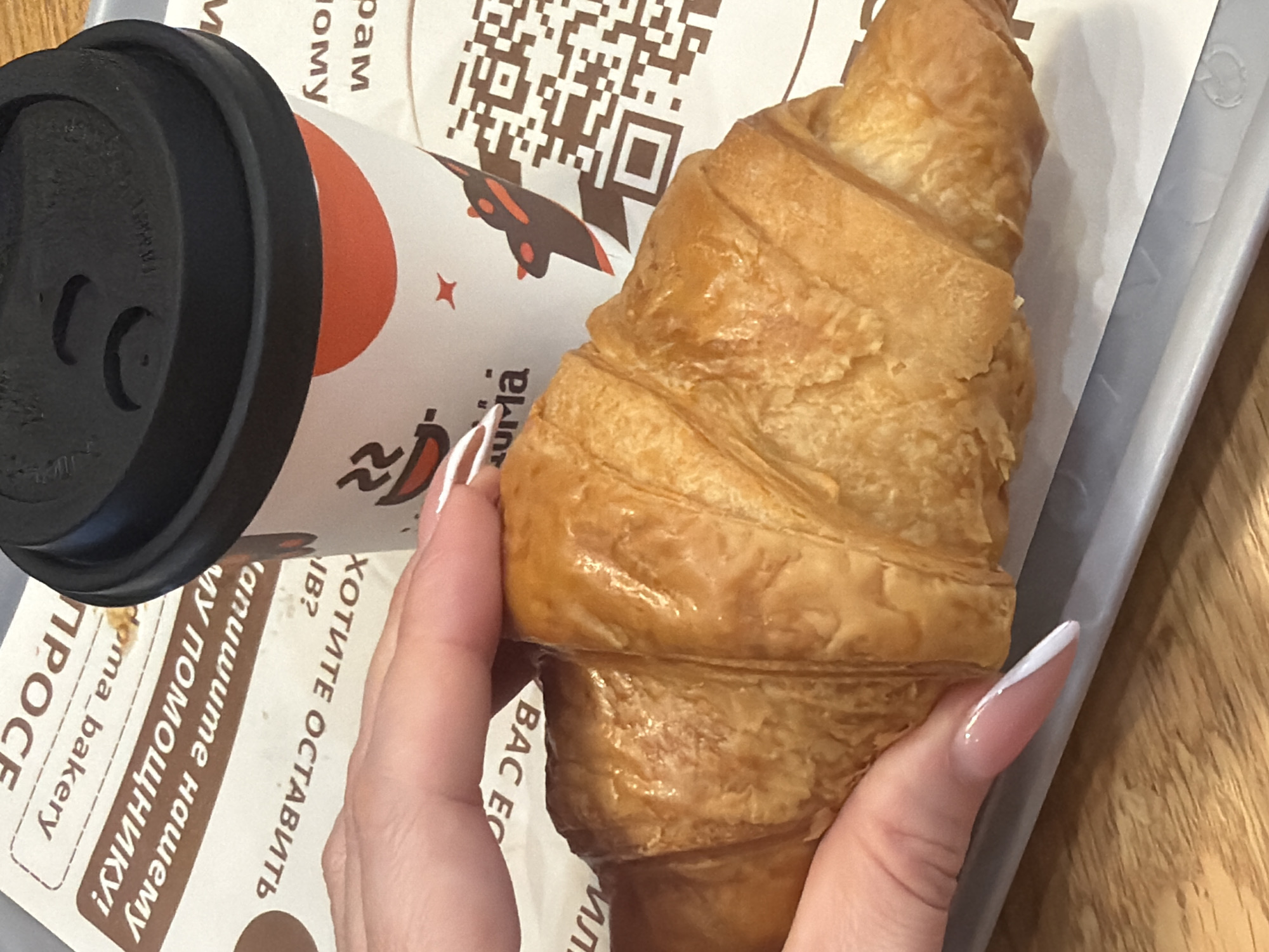 The image displays a close-up, overhead perspective of a breakfast or snack setup in an indoor cafe or bakery setting. A light-skinned hand with long, almond-shaped fingernails, featuring a pinkish-nude base and distinct white tapered tips in a French manicure style, firmly grips a large, golden-brown, glazed croissant.
To the upper right of the croissant stands a white disposable coffee cup with a textured black plastic lid. The cup is adorned with a brand logo that includes a stylized brown coffee cup with steam, the word "urma," an orange circular graphic, a small red star, and a brown rocket-like illustration, alongside a visible QR code.
Both the croissant and coffee cup are positioned on a white paper liner, which rests on a light grey, rectangular serving tray. The paper liner contains partially visible Cyrillic text printed in brown, including phrases such as "дома bakery" (home bakery), "НУ ПОМОЩНИК!" (Oh, helper!), and "хотите оставить" (Do you want to leave), along with another QR code. Beneath the tray, a light brown wooden surface, indicative of a table, is visible.
The branding elements, specifically "urma" and the Cyrillic text, identify the location as an "URMA | Coffee & Croissants" establishment in Saint Petersburg, Russia. The scene captures a singular individual's hand interacting with food and beverage items in a casual dining context.