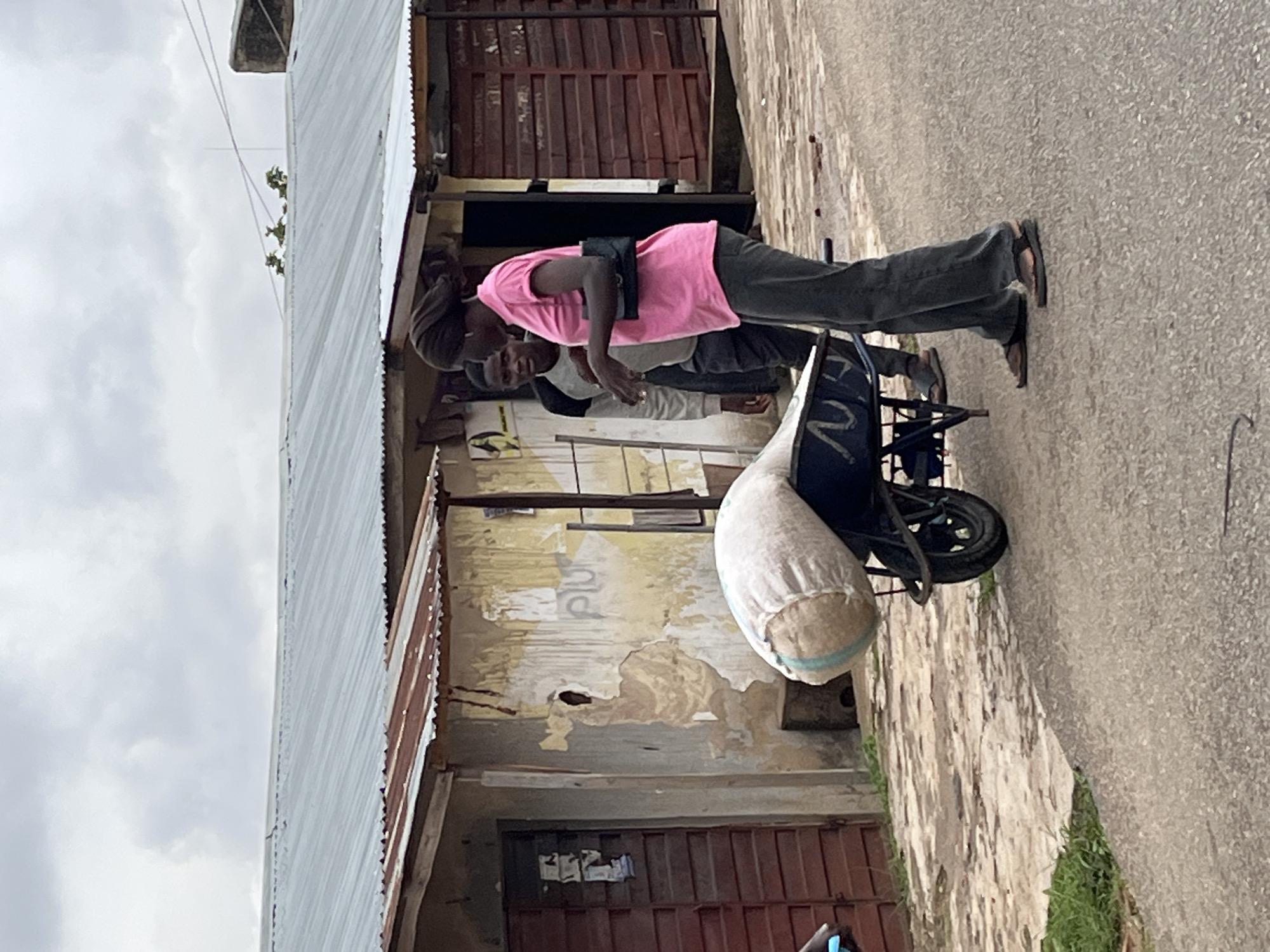 In Jos, Nigeria, a woman in a bright pink t-shirt and dark pants stands next to a wheelbarrow laden with a large, burlap sack. She is facing towards the left of the frame, looking at a younger man standing behind the wheelbarrow. The man is wearing a grey sweatshirt and dark pants, and he is looking forward. The wheelbarrow appears to be on a paved surface, possibly a street or pathway, with buildings constructed of corrugated metal roofing and rough, textured walls in the background. The sky is overcast, suggesting a cloudy day. The overall scene depicts a moment of interaction between two individuals engaged with the heavy load in the wheelbarrow.