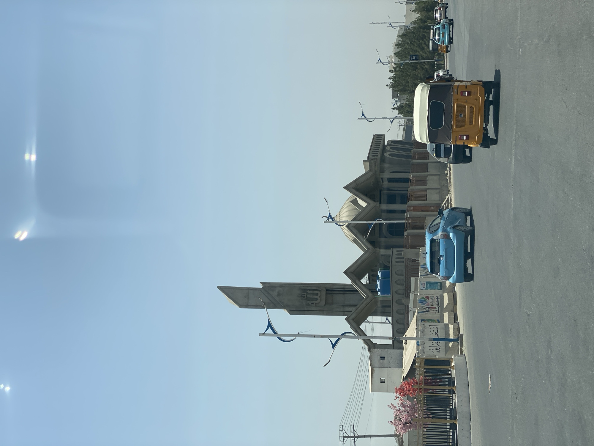 The image shows a street scene in the city of Kandahar, Afghanistan, on a clear, sunny day. The main focus is a large, unfinished concrete structure that appears to be a mosque, with a tall, angular minaret featuring Arabic calligraphy and multiple domes. In the foreground, a yellow and brown auto-rickshaw is driving away from the camera, with a blue sedan in front of it. Several other vehicles are visible on the road, including a dark sedan and a light blue car. The street is paved and appears to be a main thoroughfare. To the left of the road, a fence encloses an area with decorative pink trees and a small white building. Streetlights with blue accents are positioned along the road. The background shows more trees and buildings, suggesting an urban or semi-urban environment. The overall impression is one of ongoing development and daily life in Kandahar.