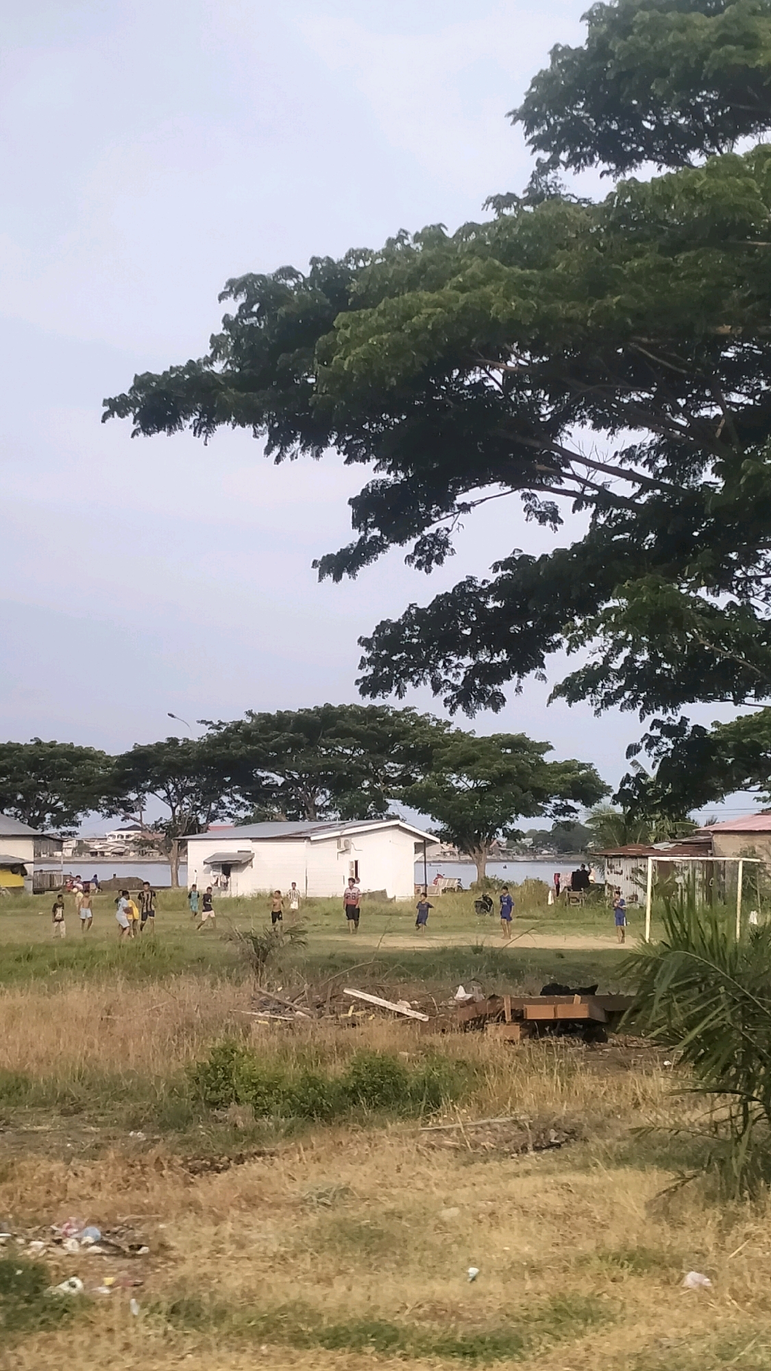 The image depicts a scene in Lhokseumawe, Indonesia, likely a public field or park area with scattered houses in the background. In the mid-ground, a group of young boys are playing soccer on a grassy field. Some are actively engaged in the game, while others are observing or moving around the field. Several small, white, rectangular buildings are visible behind the boys, suggesting a residential or community area. Beyond the buildings, a body of water, possibly a river or the sea, can be seen, with faint outlines of boats and structures on the opposite shore. Large, leafy trees with expansive canopies dominate the upper and middle sections of the image, casting shadows and contributing to the natural setting. The foreground consists of dry, overgrown grass with some scattered debris, including plastic waste. The sky is overcast, suggesting a diffused light condition.