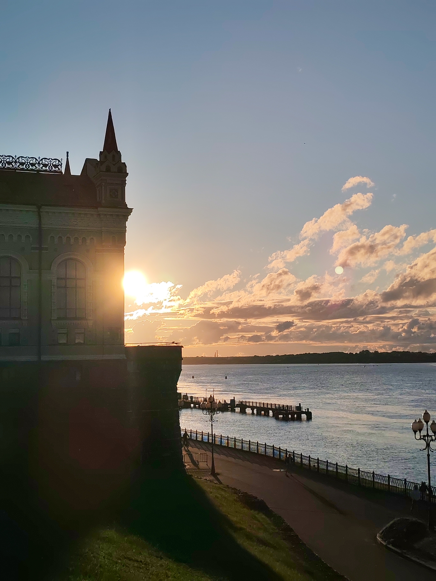 This image captures a picturesque scene at sunset in the city of Bol'shie Mkhi, Russia. The foreground is dominated by a historical building with intricate architectural details, including a pointed spire and decorative ornamentation along its roofline. The setting sun casts a warm glow, with its rays directly illuminating a section of the building, creating a lens flare effect.
Beyond the building, a wide expanse of water stretches to the horizon, reflecting the soft colors of the sky. A wooden pier juts out into the water, and further in the distance, a tree-lined shore is visible. Along the waterfront, a paved pathway runs parallel to the water, bordered by an ornate black metal fence. Several lampposts with classic designs are spaced along the pathway.
A few individuals are present, walking or standing along the pathway, adding a sense of scale and activity to the tranquil setting. The sky is a gradient of blues, with scattered clouds tinged with the warm hues of sunset. The overall atmosphere is serene and evocative of a beautiful evening by the water.
