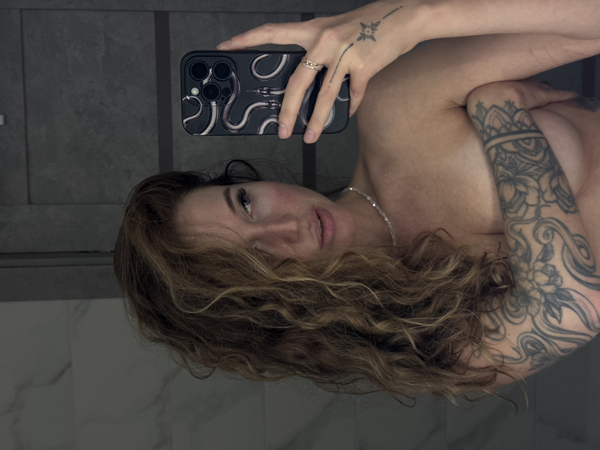 A woman with long, curly, light brown hair is taking a mirror selfie in what appears to be a bathroom. She is holding a smartphone with a snake-patterned case in her right hand, using it to capture her reflection. She is wearing a silver necklace and has a tattoo on her left arm that covers a significant portion of it, featuring floral and swirling designs. Another small tattoo is visible on the back of her right hand. Her left arm is positioned across her chest, obscuring part of her torso. The background shows tiled walls with a marble pattern and a darker, textured surface. The lighting is dim, creating shadows. There is no identifiable information about the location being Chaikovsky, Russia.