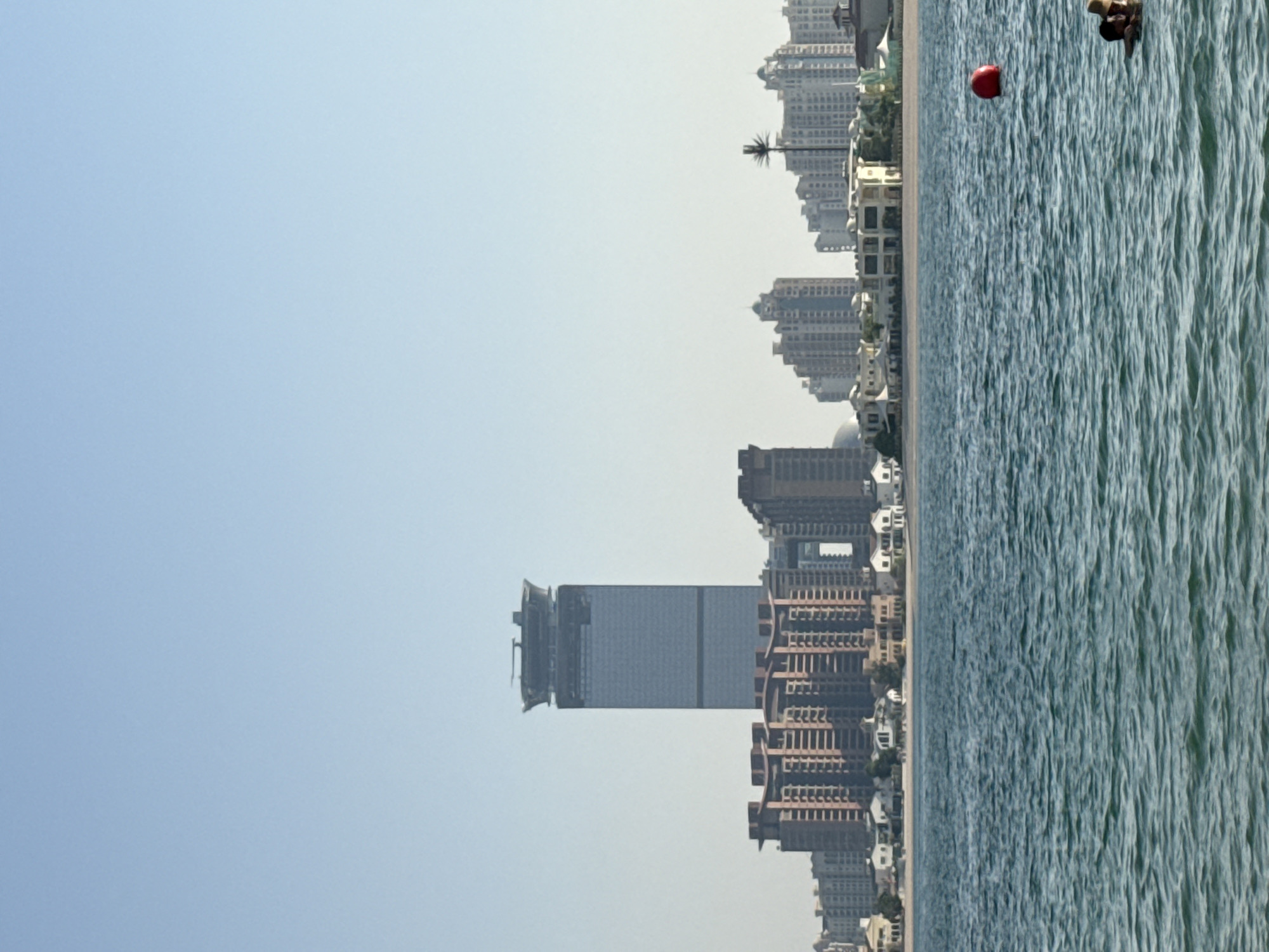 In Dubai, United Arab Emirates, a tranquil blue sea stretches across the foreground, its surface rippled by gentle waves. In the mid-ground, a cluster of buildings lines the shore, featuring a mix of modern high-rises and lower-rise residential structures. Dominating the skyline is a distinctive tall skyscraper with a broad, flat top, likely a hotel or commercial building. A red buoy floats on the water, signaling a boundary or marker. Two people are partially visible in the water on the right side of the frame, one wearing a hat and appearing to be submerged. The sky above is a clear, pale blue, suggesting a bright, sunny day.