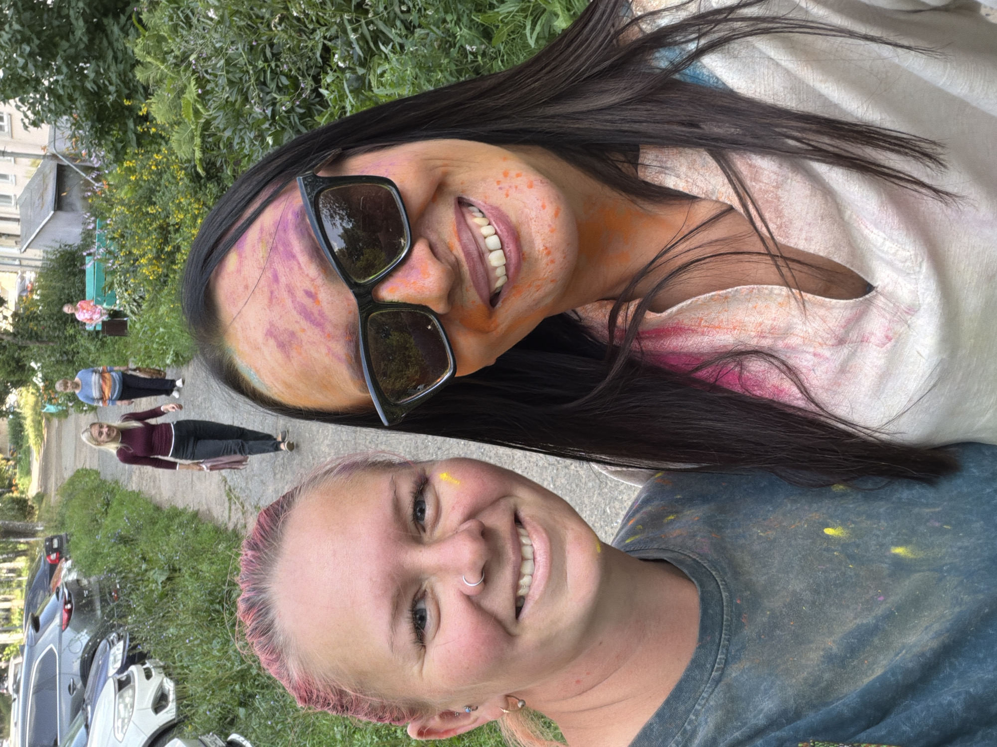 The image shows two women in the foreground, both smiling and appearing to have participated in a color festival, as evidenced by the powder paint on their faces and clothing. The woman on the left has pink-dyed hair, a nose ring, and is wearing a dark t-shirt. The woman on the right has long dark hair, sunglasses, and is wearing a light-colored shirt. In the background, a pathway is visible with parked cars on the left and lush greenery on the right. Further down the path, two more women are walking, one in a burgundy top and dark pants, and another in a striped sweater. In the far background, a residential building can be seen. The overall scene suggests an outdoor gathering or event in a semi-urban or suburban environment. The presence of parked cars and a residential building points to a location within a city or town. Based on the context provided, this scene is located in the city of Chaikovsky, Russia.
