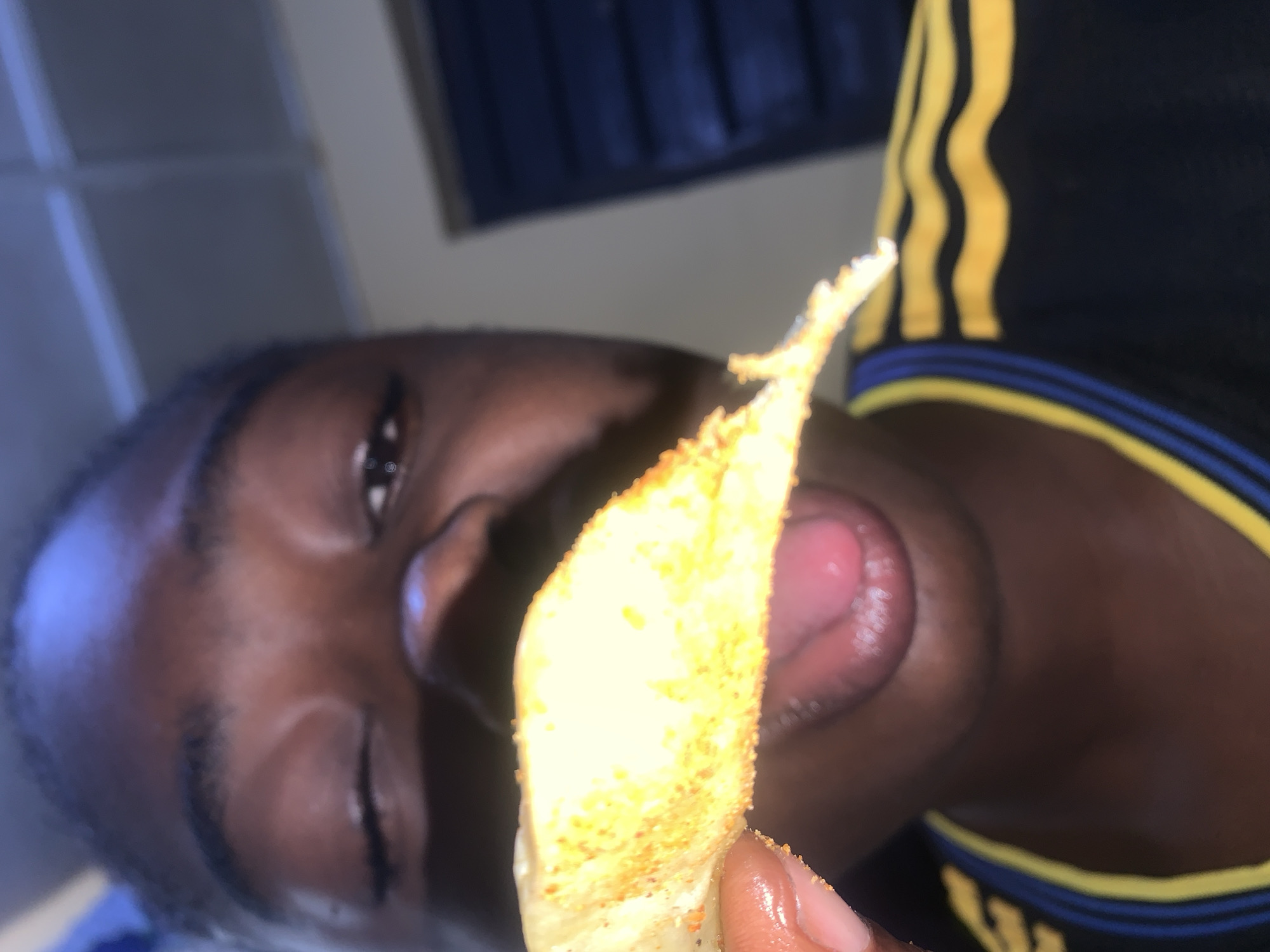 The image shows a close-up of a person's face as they are about to eat a piece of seasoned food, possibly a chip or cracker. The person is winking, and their tongue is slightly extended. The food item is golden brown and appears to be coated in spices. The person is wearing a dark t-shirt with yellow and blue stripes on the collar. The background is blurry, but suggests an indoor setting with a blue shuttered window. The image quality is moderate. No specific location in Rafin Par, Nigeria can be determined from the image itself.
