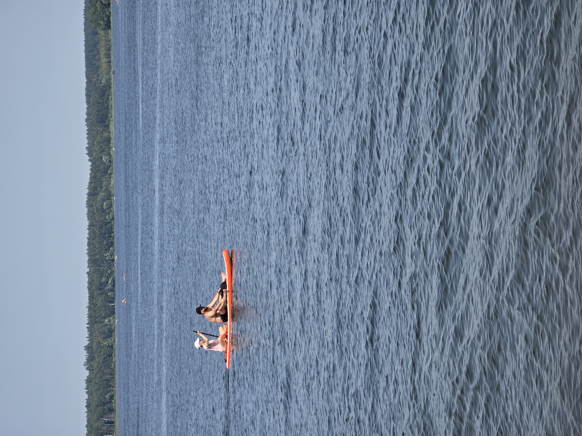 Two people are paddleboarding on a lake. A woman wearing a pink shirt and white hat is in the front, holding a paddle. A man with a dark beard and black baseball cap is behind her, also holding a paddle. They are on an orange paddleboard. The lake is calm with small ripples on the surface. In the background, a dense green forest lines the shore of the lake. The sky is a clear, pale blue. The scene is set in the city of posyolok Gidrouzla imeni Kuybysheva, Russia.