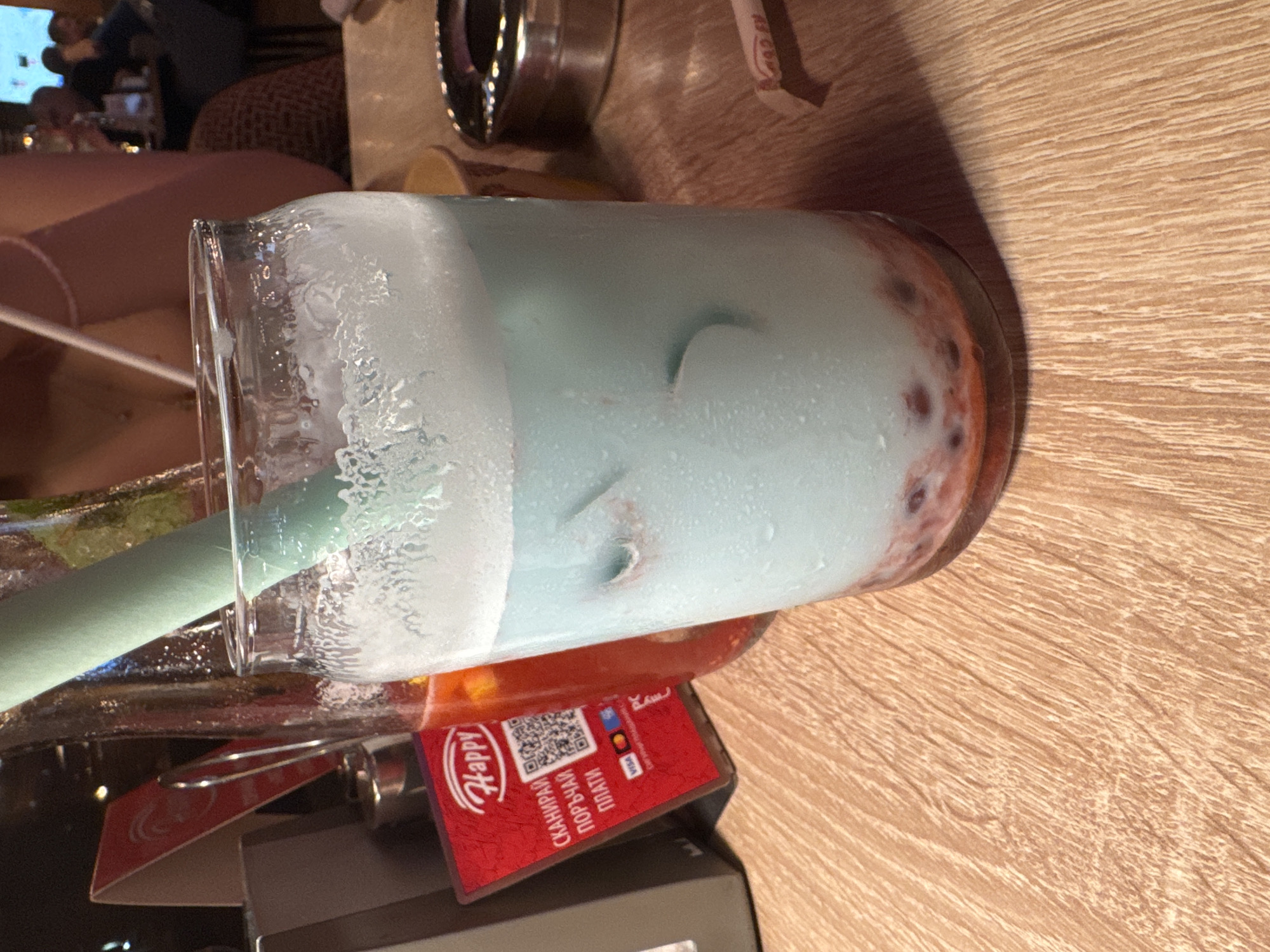 The image displays a refreshing light blue beverage in a tall, clear glass, positioned on a light wooden table with a visible grain. The drink features a thick, white, foamy layer on top and exhibits condensation on the exterior of the glass, indicating its cold temperature. At the bottom of the glass, small, reddish, spherical elements, likely boba pearls or fruit pulp, are visible. A light green straw is inserted into the drink.
The scene is set indoors, likely in a cafe, bar, or restaurant, suggested by the table, subtle background lighting, and blurry figures in the distance. The ambient light implies it is evening or nighttime.
A red payment card or stand is partially visible near the base of the glass. It features a "Happy t" logo, a QR code, and Cyrillic text that translates to "SCAN, ORDER, PAY." Visa and Mastercard logos are also present on the card. An ashtray is partially visible in the upper background. The overall impression is one of a casual, relaxed indoor setting in Sofia, Bulgaria.