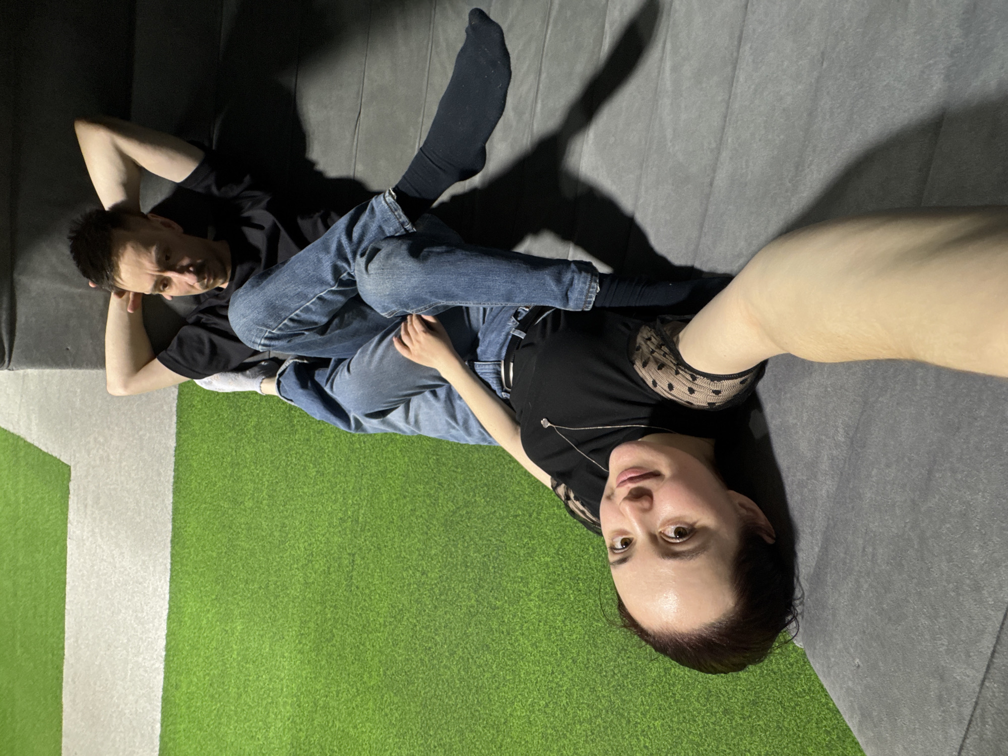 This full-shot selfie captures a man and a woman relaxing on the floor of what appears to be an indoor recreational facility in Izhevsk, Russia. The image is taken from a slightly elevated angle, with the woman holding the camera.
The woman, positioned in the foreground, lies on her back, looking directly at the camera with a calm, neutral expression. She has dark hair, brown eyes, and is wearing a black top with delicate, sheer, polka-dotted lace detailing on the shoulder. A silver necklace with a small pendant adorns her neck. Her legs are stretched out, and she wears white socks.
Behind and to the left of the woman, the man is also lying on his back, with his head resting on his clasped hands. He looks slightly away from the camera, his expression relaxed. He has short, dark hair, a black t-shirt, and blue jeans. His legs are crossed, with one leg resting over the woman's lower legs. He wears black socks.
The floor is a mix of surfaces: a soft, grey padded area where the man is mostly situated, and a bright green, short-pile turf-like material where the woman's lower body and arm extend. A clean white line demarcates sections of the green surface, suggesting a designated play or activity zone. The environment appears dimly lit, possibly with artificial light, casting distinct, elongated shadows of the subjects onto the grey surface behind them. There is no visible text in the image.
The overall atmosphere is one of casual relaxation and companionship, suggesting the couple is taking a break during an outing to a fun, active indoor venue.