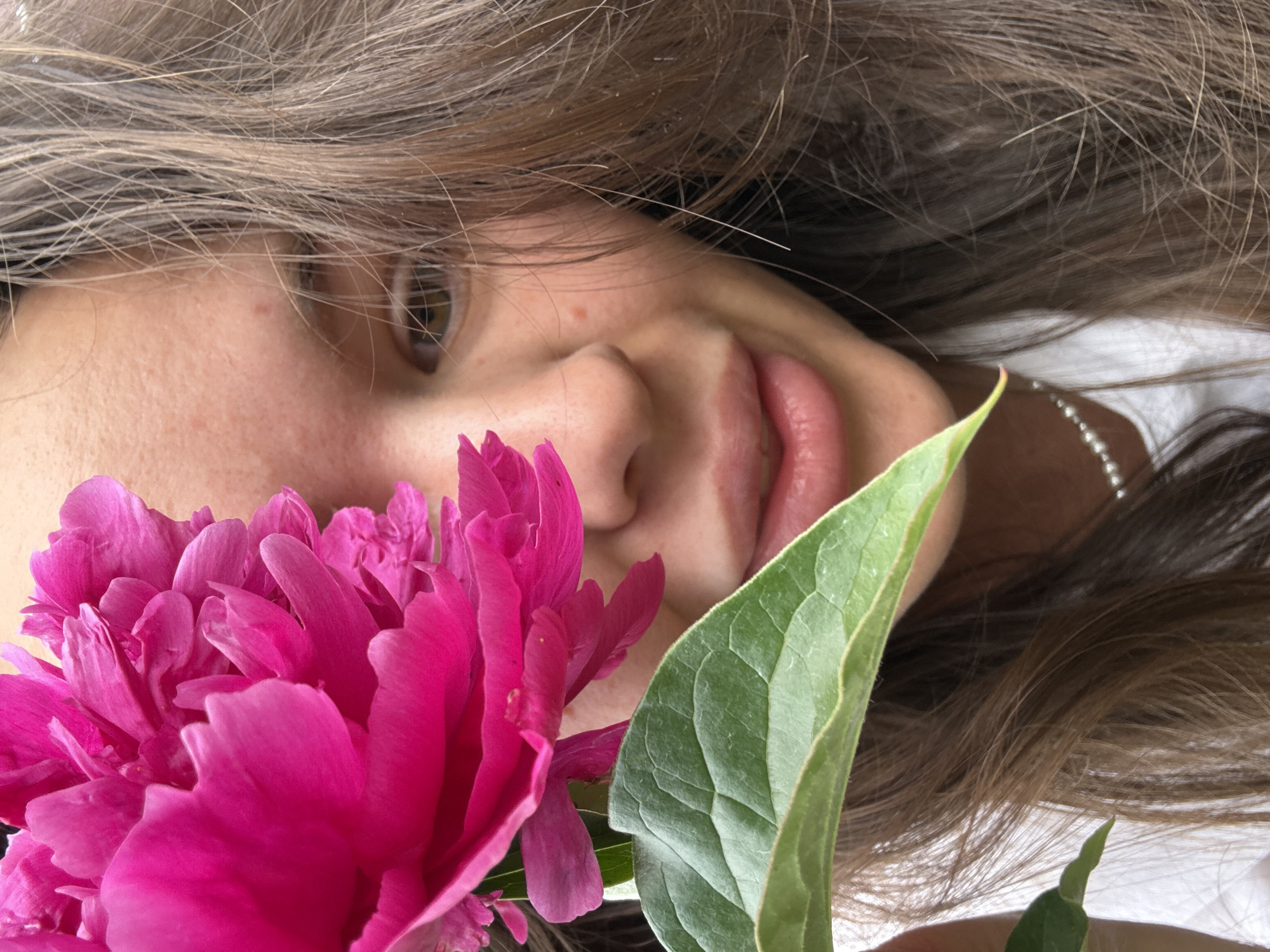 A close-up, eye-level, medium shot features a young woman partially obscured by a vibrant pink peony flower and a large green leaf in the foreground. The woman's face is turned slightly to the right, with her left eye visible and her right eye hidden by the flower. Her lips are parted as if speaking or smiling. She has long, brown, slightly wavy hair that frames her face and extends down her shoulders. A string of small pearls is visible around her neck. The background is blurred, suggesting an outdoor setting, possibly a garden or natural environment. The lighting is soft and natural, highlighting the textures of the flower, leaf, and skin. No specific location in Bely Yar, Russia, can be identified from the image.