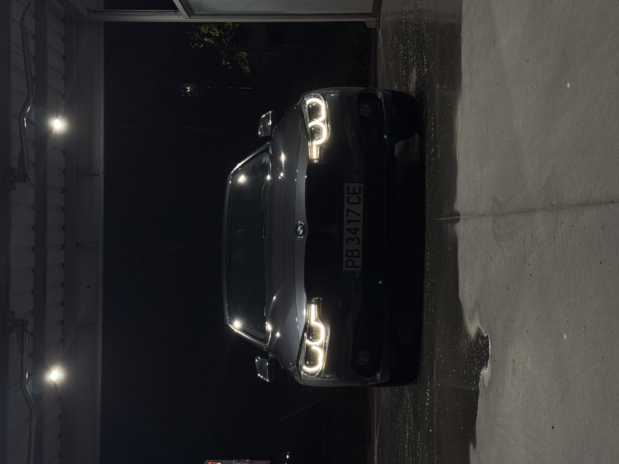 The image shows a dark gray BMW car parked inside a garage or car wash facility. The license plate reads "PB 3417 CE," indicating it is registered in Bulgaria. The headlights are on, illuminating the front of the vehicle. To the left of the car is a wall with industrial lighting. To the right is a wet concrete floor. The environment suggests an indoor, utilitarian space. Without additional context, the exact location in Plovdiv, Bulgaria, is undetermined.