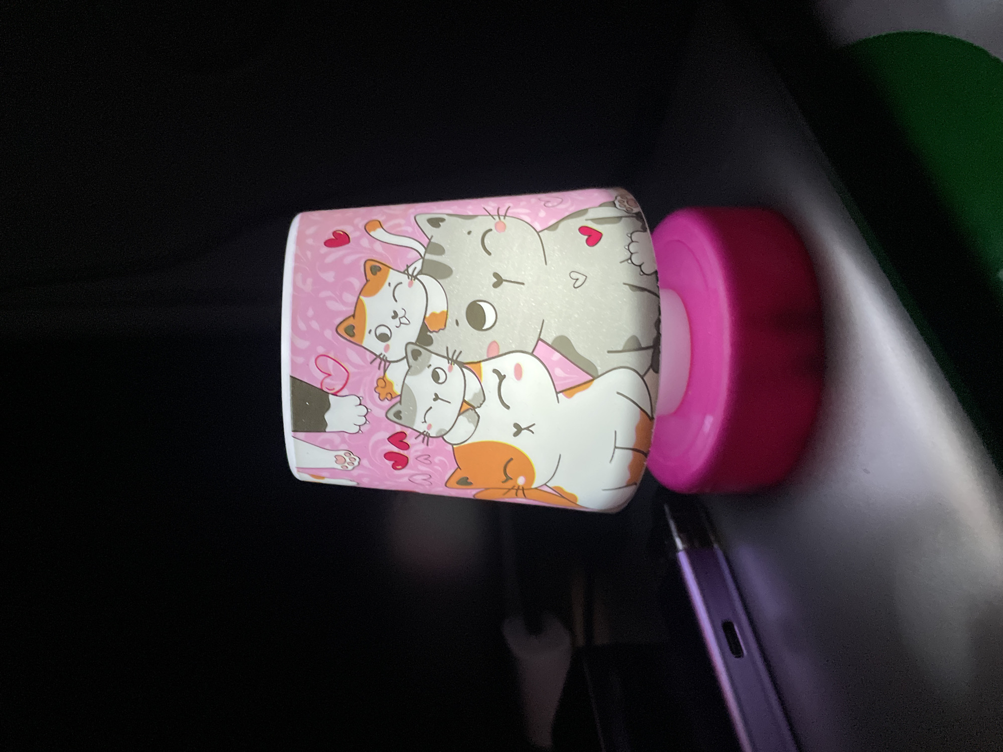 A pink, cylindrical night light with a pattern of cartoon cats sits on a dark, reflective surface. The lamp is illuminated from within, casting a soft glow on the surrounding area. To the left of the lamp, a portion of a dark, rectangular object, possibly a phone or tablet, is visible, reflecting the light. The background is dark and indistinct. There is no indication of a specific location within Saint Petersburg, Russia.