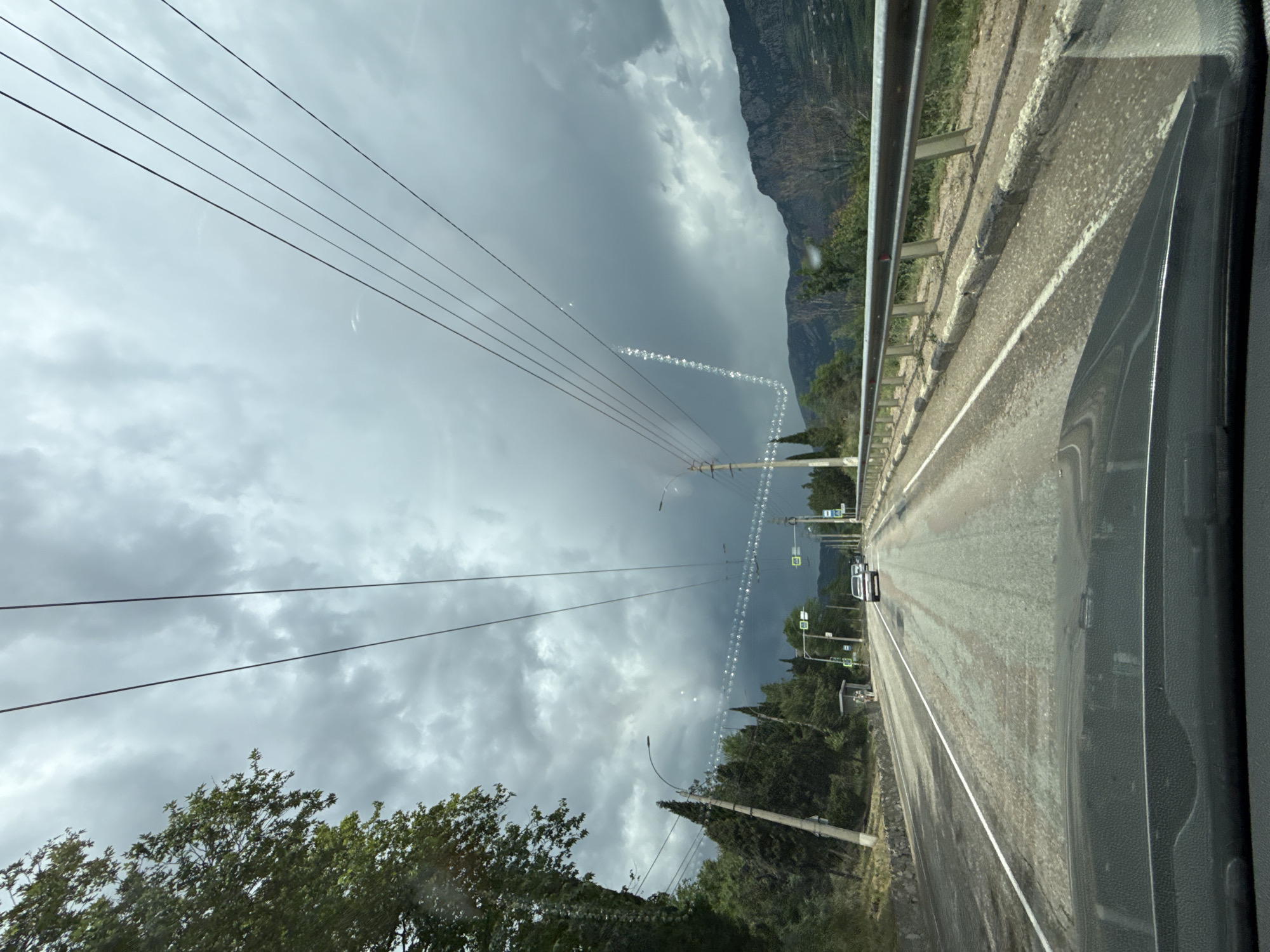 An overcast, gray sky looms over a road bordered by trees and mountains. The perspective is from inside a vehicle, looking forward through the windshield. Power lines stretch across the upper portion of the frame. Several cars are visible on the asphalt road. The road has a white line marking its center. On the right side of the road, a metal guardrail runs alongside. On the left, a stone wall and trees line the road. The overall scene suggests an outdoor, possibly rural, environment under cloudy conditions.
