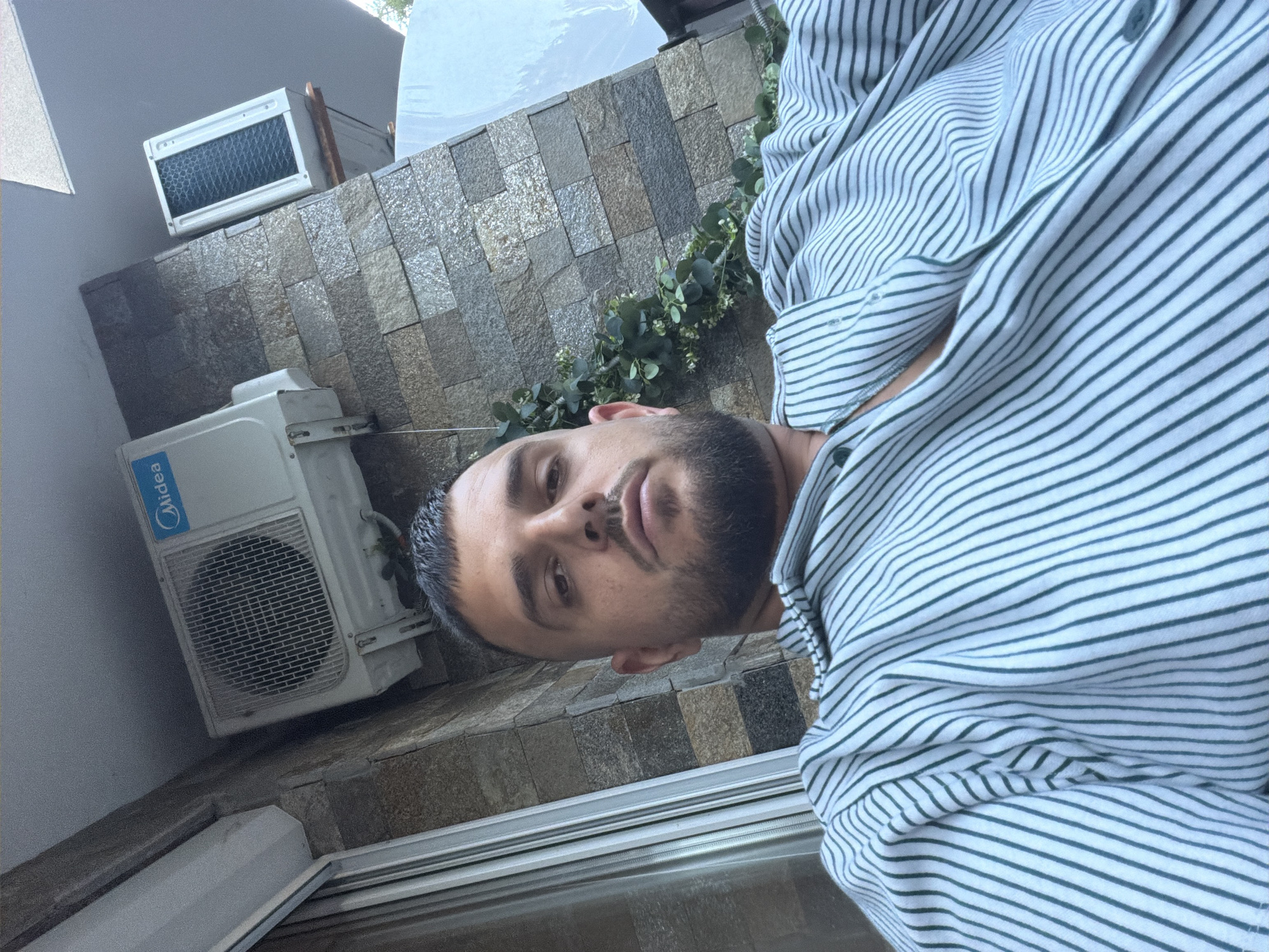 A man is taking a selfie outdoors, seated in front of a stone-tiled wall. He has short dark hair and a beard. He is wearing a collared shirt with green and white stripes. Two air conditioning units are visible on the wall behind him. One is a Midea brand unit, and the other is a white unit with a black filter. Green foliage is draped on the wall between the two units. A window with blinds is visible to the left of the frame. The scene appears to be on a balcony or patio area. This location is identified as being in Nesebar, Bulgaria.