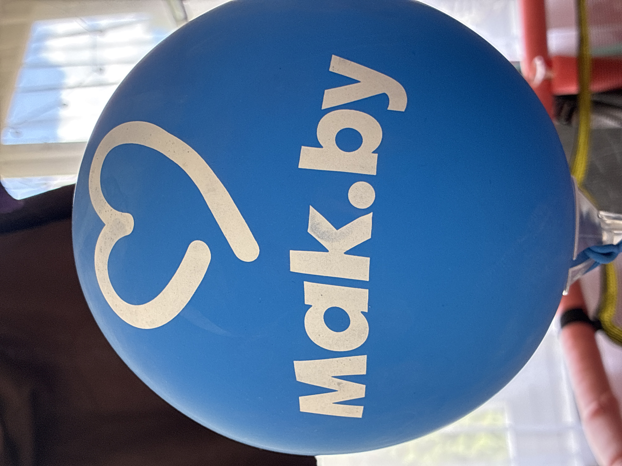 A close-up, slightly upward angled shot shows a bright blue balloon with a white outline of a heart at the top and the text "Mak.by" in white letters below it. The balloon is the primary focus, set against a softly blurred background that includes a dark, out-of-focus area to the left and a brightly lit window with white curtains and a hint of blue sky and clouds to the right. Below the balloon, parts of a red object and a yellow strap are visible, suggesting the balloon is being held or displayed. The lighting is natural, coming from the window. No specific details about the location in Гаранскі сельскі Савет, Belarus are discernible from the image itself; the scene is generic and could be anywhere.