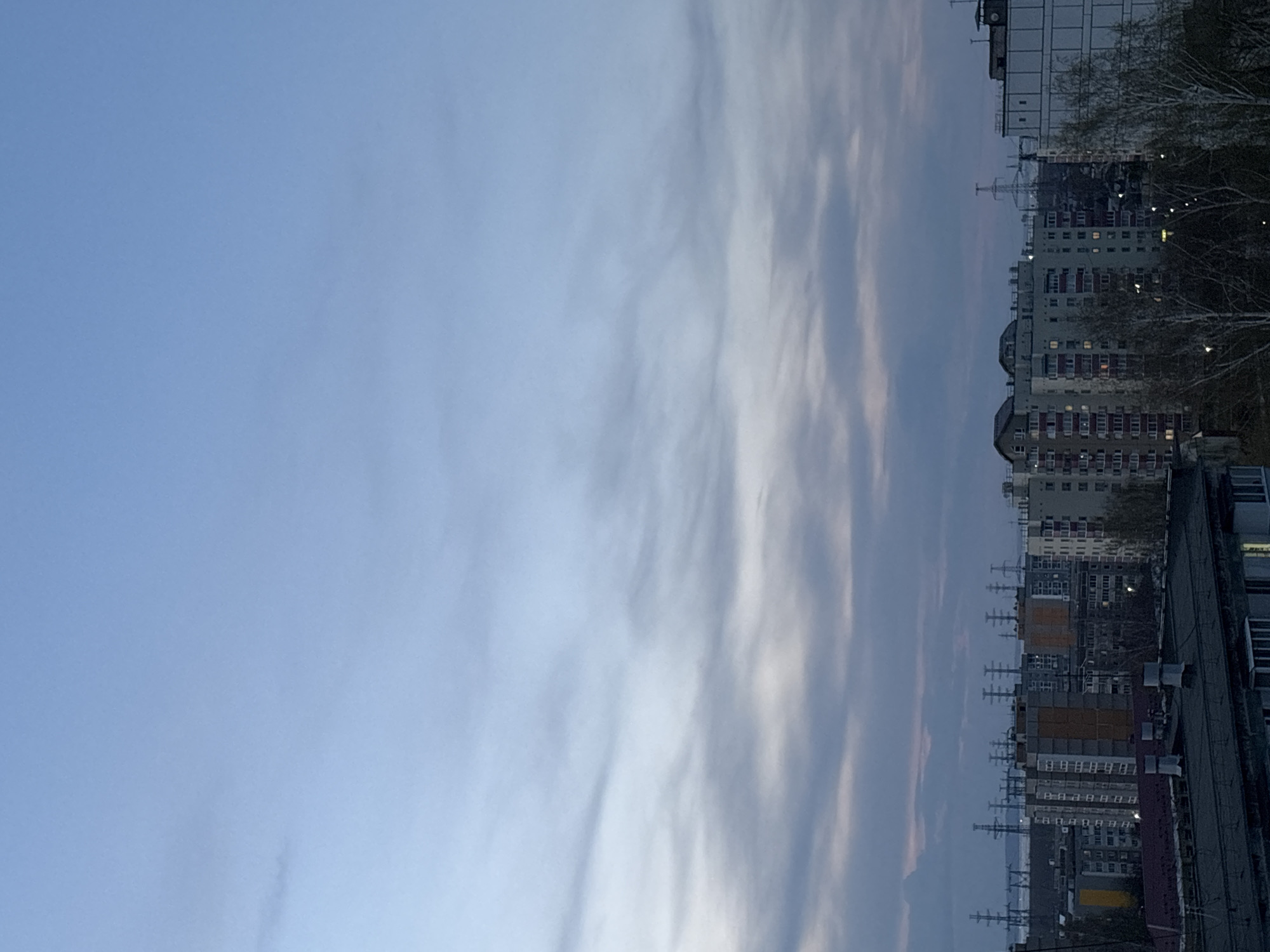 The image shows a cityscape in Chaikovsky, Russia, at dusk. The sky is a gradient of light blue to pale pink, with long streaks of clouds. In the foreground are rows of apartment buildings of various heights and colors, constructed from brick and concrete. Power lines run above the buildings. Some windows are lit, suggesting residents are home. Bare trees are interspersed amongst the buildings.