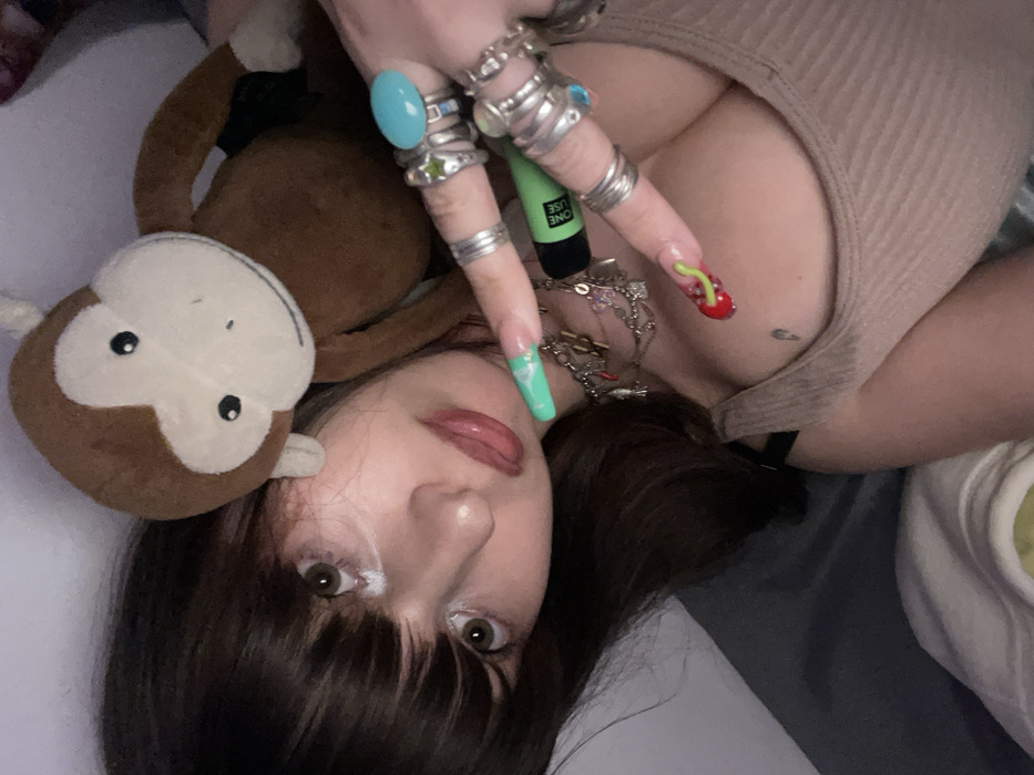 A young woman is posing while lying down. She has brown hair, fair skin, and brown eyes. She's wearing multiple rings and a neutral-colored tank top. A stuffed monkey is to her left, resting on the surface she is lying on. Her nails are long and decorated. She is holding a green vape pen. A silver necklace adorns her neck. Her eye makeup features white eyeshadow. The overall composition is casual and intimate.