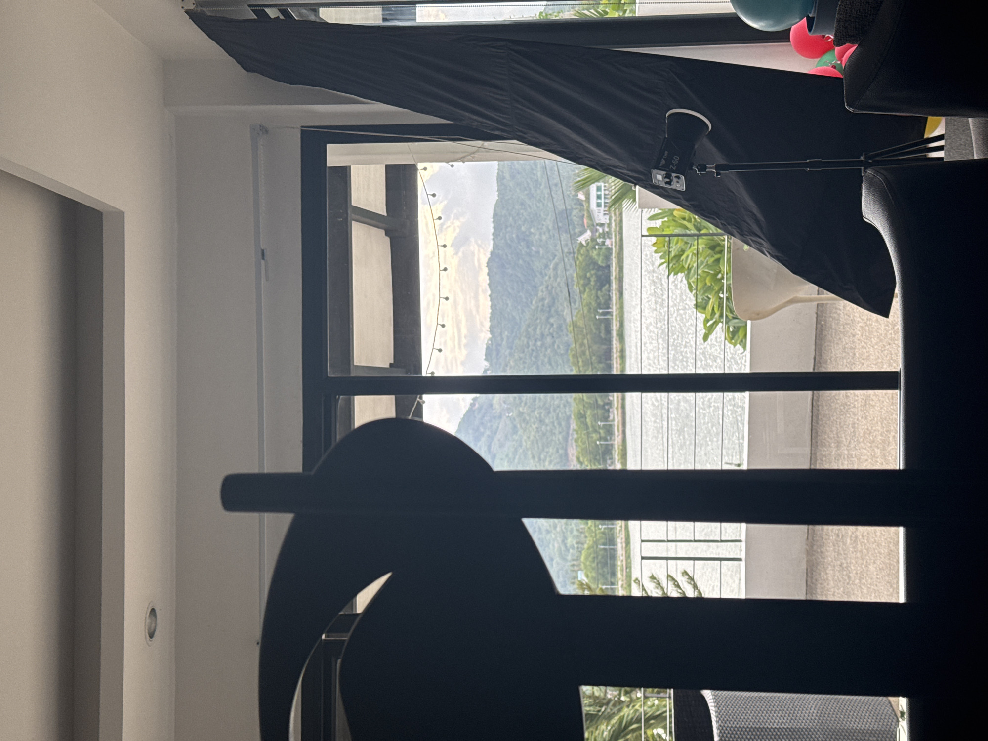 The image shows a view from inside a building looking out through a large window or glass door. In the foreground, a dark silhouette of a decorative object partially obstructs the view. Beyond this, a balcony with a white railing is visible, leading to a body of water that appears to be a lake or calm sea. Lush green hills and mountains form the background, with some buildings scattered amongst the trees on the slopes. The sky above is overcast with hints of sunlight. On the balcony, a black photography umbrella is set up on a tripod, with a light fixture attached. There are also some balloons visible in the lower right corner of the image, suggesting a festive occasion. The overall impression is of a scenic view from a comfortable indoor space, likely a home or a resort, possibly in a tropical location like Ko Samui District, Thailand, given the vegetation and the overall aesthetic.