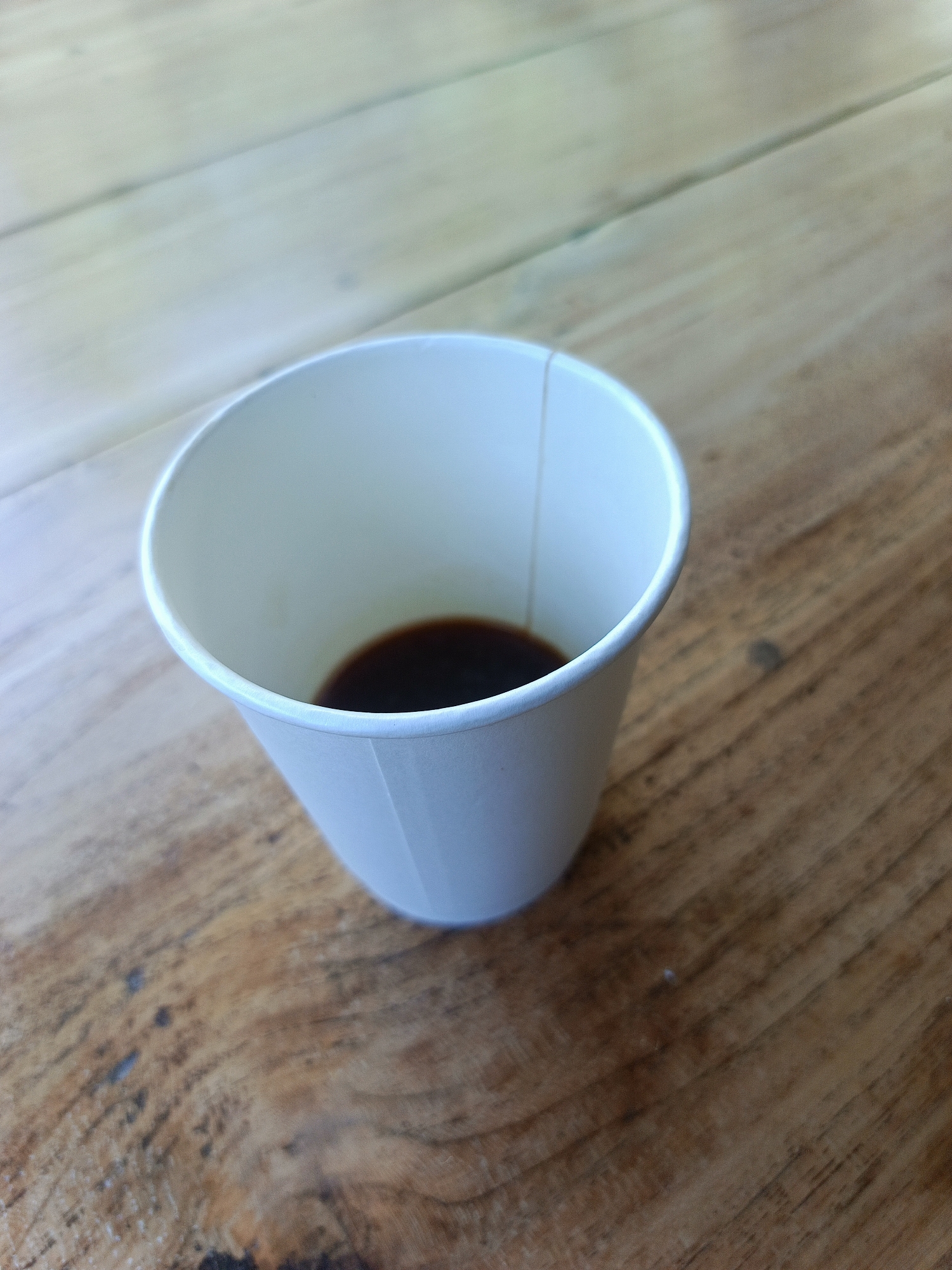 A white paper cup containing a small amount of dark liquid sits on a wooden surface. The cup is positioned slightly to the left of the frame's center, with its opening facing upwards. The liquid is visible at the bottom of the cup. The wooden surface is made of planks with visible grain and some imperfections, suggesting an outdoor or rustic setting. The lighting indicates it is daytime, with soft shadows cast by the cup. The background shows more of the wooden surface, with diagonal lines formed by the grain of the wood. The image is taken from a slightly elevated angle, looking down at the cup. The scene is devoid of any human presence or other objects. The location in Kuta, Indonesia, is not determinable from the visual information provided.