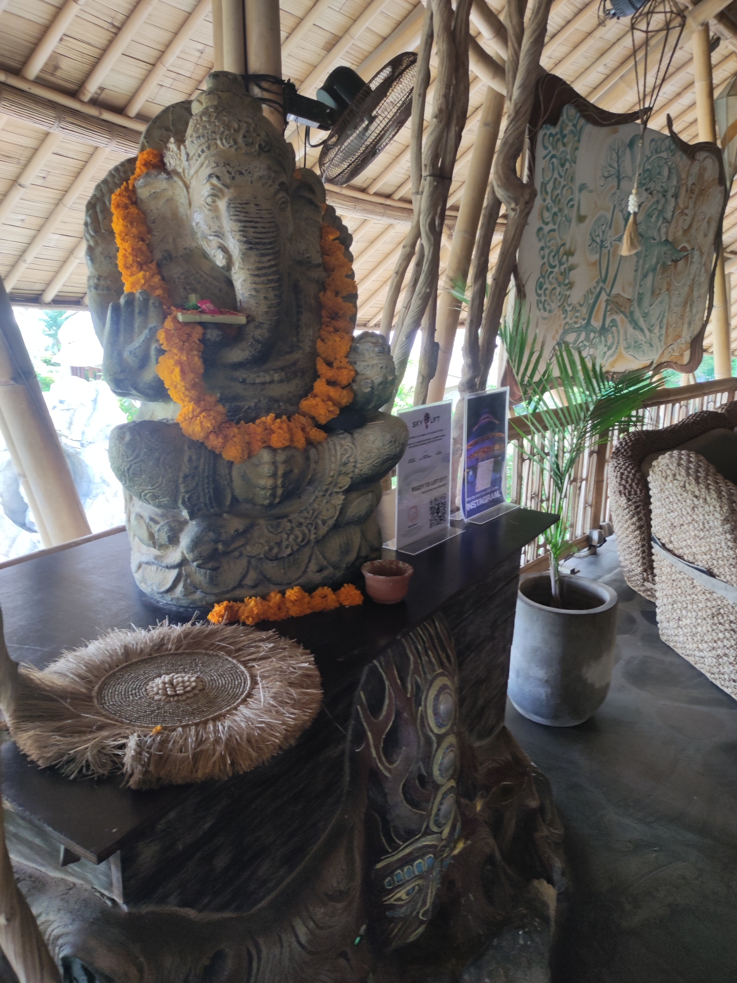 The image depicts an indoor scene with a prominent stone statue of the Hindu deity Ganesha adorned with an orange garland. The statue rests on a dark, intricately carved wooden structure. A decorative woven mat and a small ceramic bowl are placed near the statue. Behind the statue, there are two clear plastic stands displaying promotional materials with the words "SKY LIFT" and "INSTAGRAM." A potted palm tree is positioned next to the wooden structure. The background reveals a bamboo structure forming the roof and walls of the building. A decorative painting hangs on one of the bamboo walls. A wicker couch is visible in the background, indicating a seating area. Based on the promotional material for SKY LIFT, the location is likely in or near a tourist attraction. The specific city in Indonesia is Unknown.