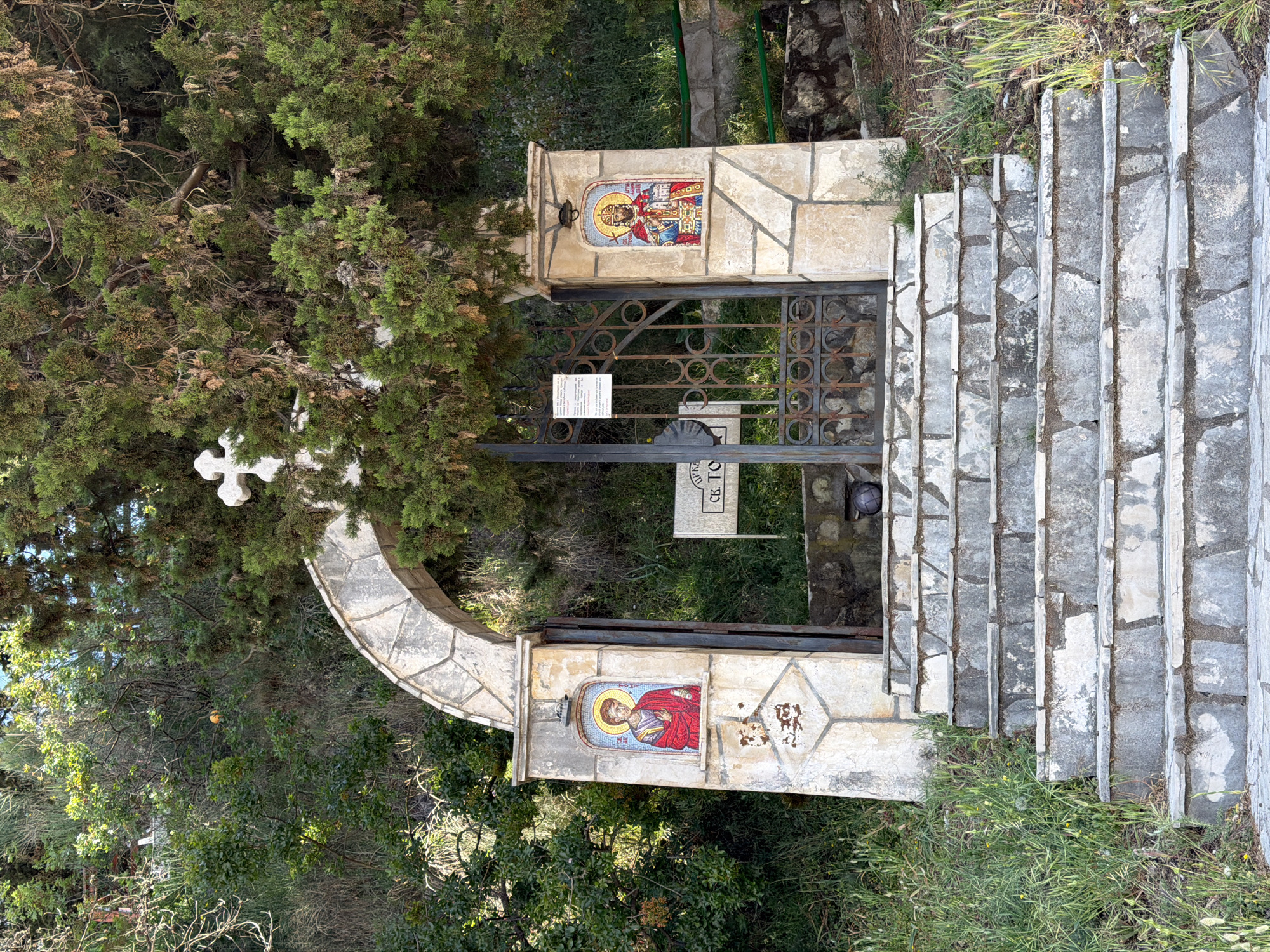 The image shows the entrance to a church or monastery in Boreti, Montenegro. A stone archway with religious mosaics on either side surrounds a metal gate. A white cross is affixed to a tree near the archway. Stone steps lead up to the gate. The area surrounding the entrance is lush with greenery.