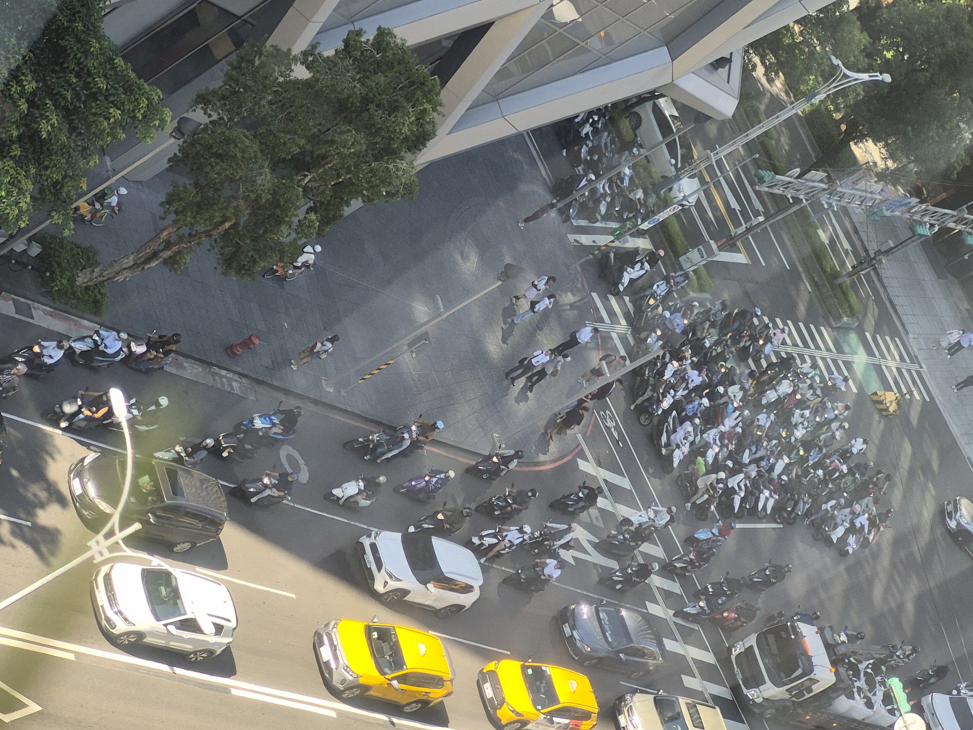 This is a high-angle, eye-level, aerial shot of a busy street intersection in Taipei, Taiwan. The image depicts a dynamic scene with a multitude of motorcycles, scooters, and a few cars navigating the urban landscape. The street is paved and marked with white lines indicating traffic lanes and pedestrian crossings.
In the foreground, several individuals are on motorcycles and scooters, some stopped at what appears to be a traffic light or intersection, while others are in motion. The vehicles are varied in color and style. Among the people, some are wearing helmets, as is common in Taiwan. A few pedestrians are also visible on the sidewalks or crossing the street.
The surrounding environment includes modern buildings with glass facades, indicative of a commercial or business district. Lush green trees line the sidewalks, providing shade and softening the urban environment. Streetlights and traffic signals are also present. The lighting suggests it is daytime, with shadows cast by the buildings and vehicles, indicating the sun's position. The overall impression is one of a bustling city, characteristic of Taipei, with a high volume of two-wheeled vehicle traffic.