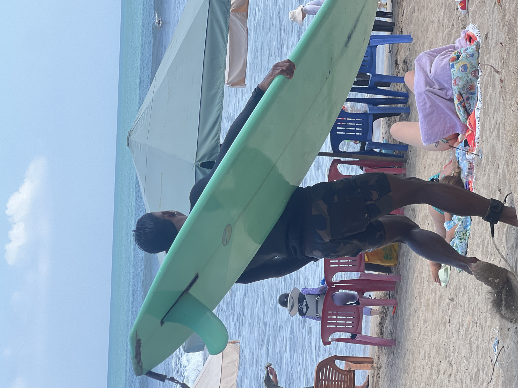 A medium shot captures a person in dark camouflage shorts and a black long-sleeved shirt carrying a large, pale green surfboard horizontally across their body. The surfboard's fin is visible on the left. The person is walking on a sandy beach, with a dust cloud trailing from their right foot as they move. In the background, the turquoise ocean stretches to the horizon under a pale blue sky with sparse clouds. Several beach chairs, including red and blue plastic ones, are scattered on the sand. A person in a purple rash guard and a white hat sits in the water, facing away from the camera. Further back, two large beach umbrellas provide shade. On the sand in the foreground, a person lies on a colorful towel, covered partially by a purple towel. The overall scene suggests a beach activity, likely surfing, in a sunny, coastal environment.