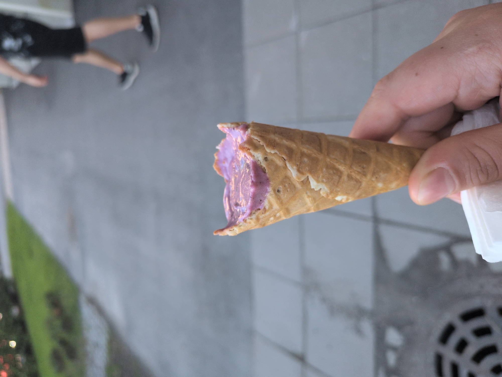 A fair-skinned hand, observed from a first-person perspective, holds a partially eaten ice cream cone in an outdoor urban setting in Helsinki, Finland. The light brown waffle cone contains melted, purple-hued ice cream or sorbet. A small white paper napkin is held beneath the cone by the same hand. The immediate foreground consists of light grey, square-tiled pavement, with a circular metal drain cover featuring a radiating spoke pattern visible in the lower-left corner. To the left of the paved walkway, a strip of green grass and blurred foliage extends. In the soft-focused background, another paved path is visible, with a blurry figure of a person walking away from the viewer, clad in dark shorts, a dark shirt, and dark sneakers. The ambient light suggests daylight, possibly late afternoon or early evening.