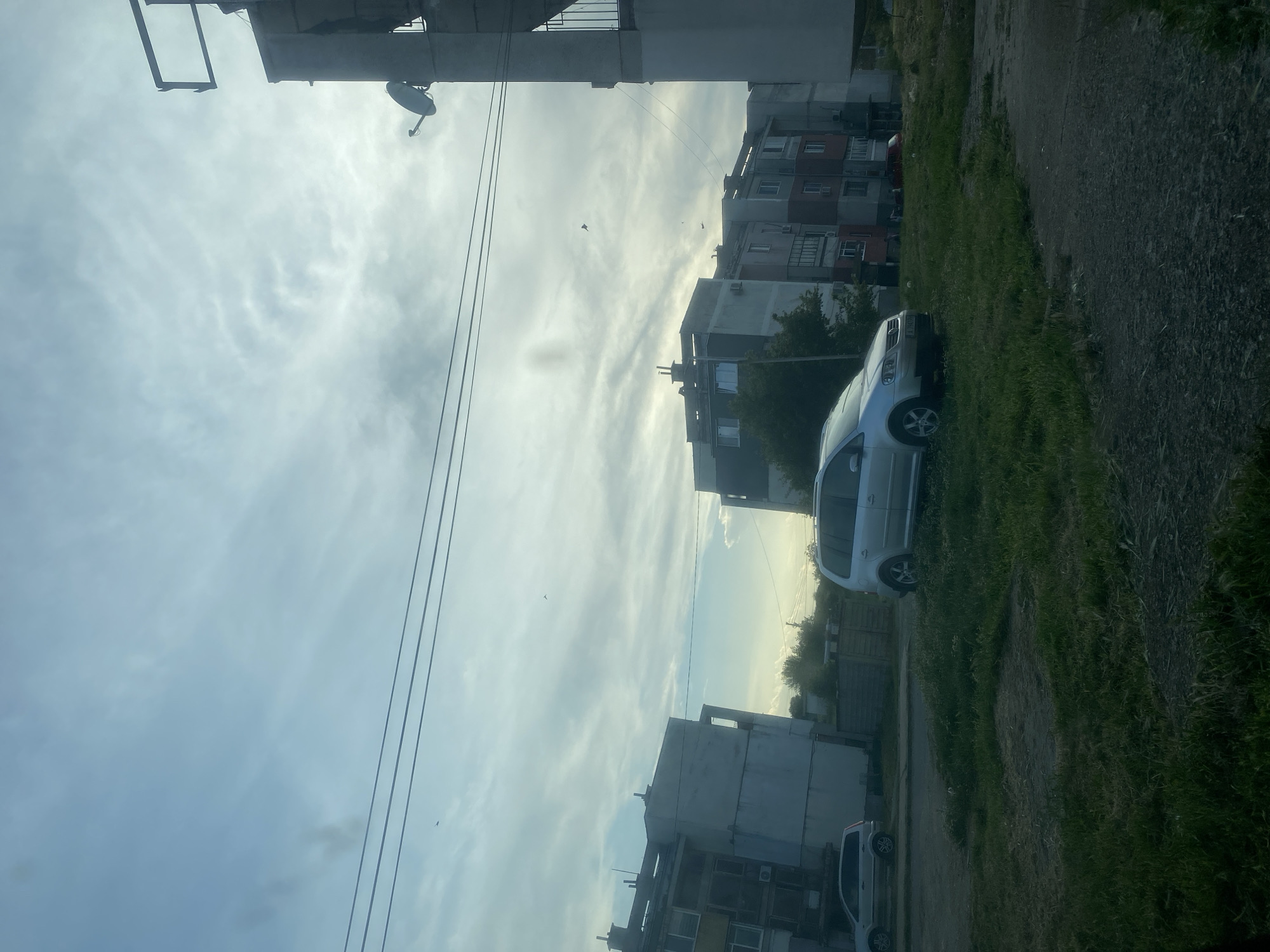 The image shows a street scene in Debelt, Bulgaria. Several multi-story apartment buildings are visible on either side of the road. A light-colored car is parked on the grassy shoulder, angled slightly onto the paved section. Power lines stretch across the sky. The sky is cloudy with patches of sunlight breaking through. The vegetation is green and unkempt. The road surface appears cracked and uneven. There is no visible human activity in the scene.
