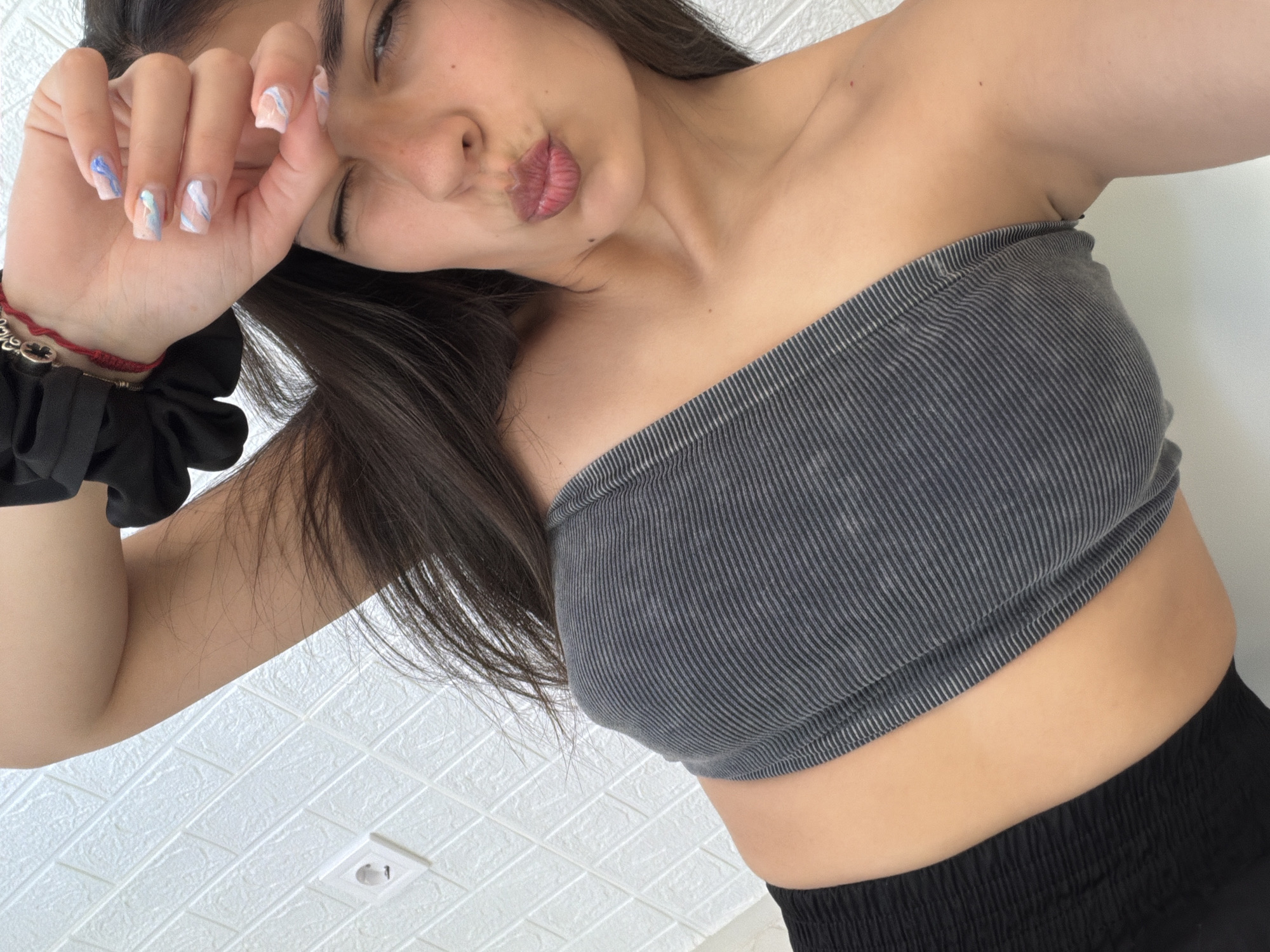 A young woman is captured in a close-up, diagonally oriented selfie, likely taken indoors in Gabrovo, Bulgaria. She is looking downwards towards the camera with her eyes partially closed or squinting, and her lips are puckered in a playful pout. Her long, dark hair cascades over her shoulders. She wears a grey, vertically ribbed tube top that exposes her midriff, paired with dark (likely black) bottoms. Her left hand is raised towards her face, showcasing her fingernails, which are painted with a light base and abstract blue and white swirl designs. On her left wrist, she has a red string bracelet adorned with silver accents and a black scrunchie. The background features a white textured surface, resembling patterned ceiling tiles or wall paneling, with a visible white electrical outlet in the lower right corner. The lighting appears bright and even, consistent with an indoor setting. The overall impression is casual and relaxed. There is no visible text in the image.
