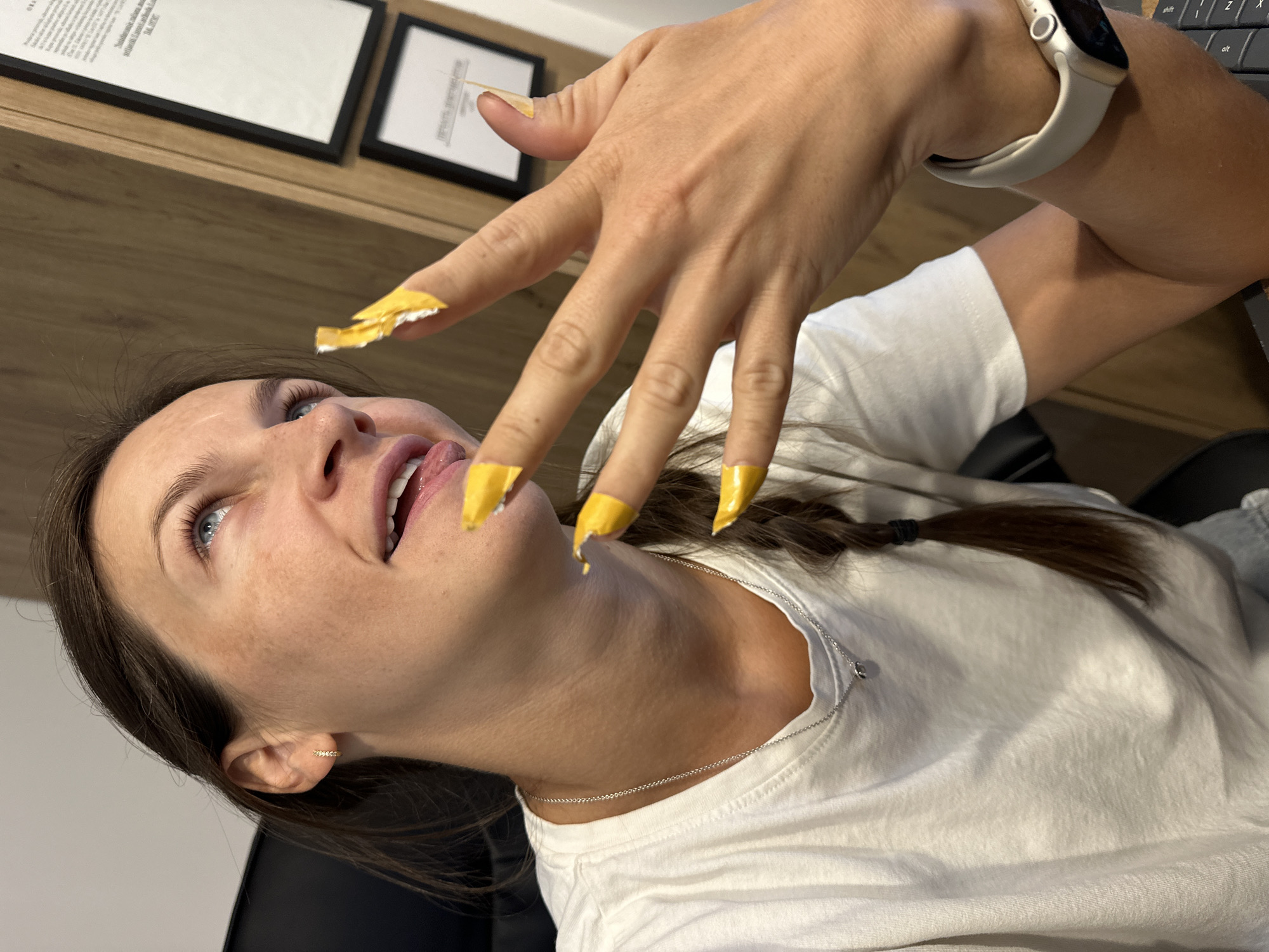 A young woman with brown hair and blue eyes is sitting in an office, looking up and to the right with her tongue sticking out slightly. She is wearing a white t-shirt and a silver necklace. Her nails are long and painted bright yellow. Her left hand is raised to her face, with her fingers extended. Behind her, there is a wooden desk and a framed document on the wall. The scene is well-lit, and the focus is on the woman and her hand. There is no indication of the city of Boreti, Montenegro, in the image.