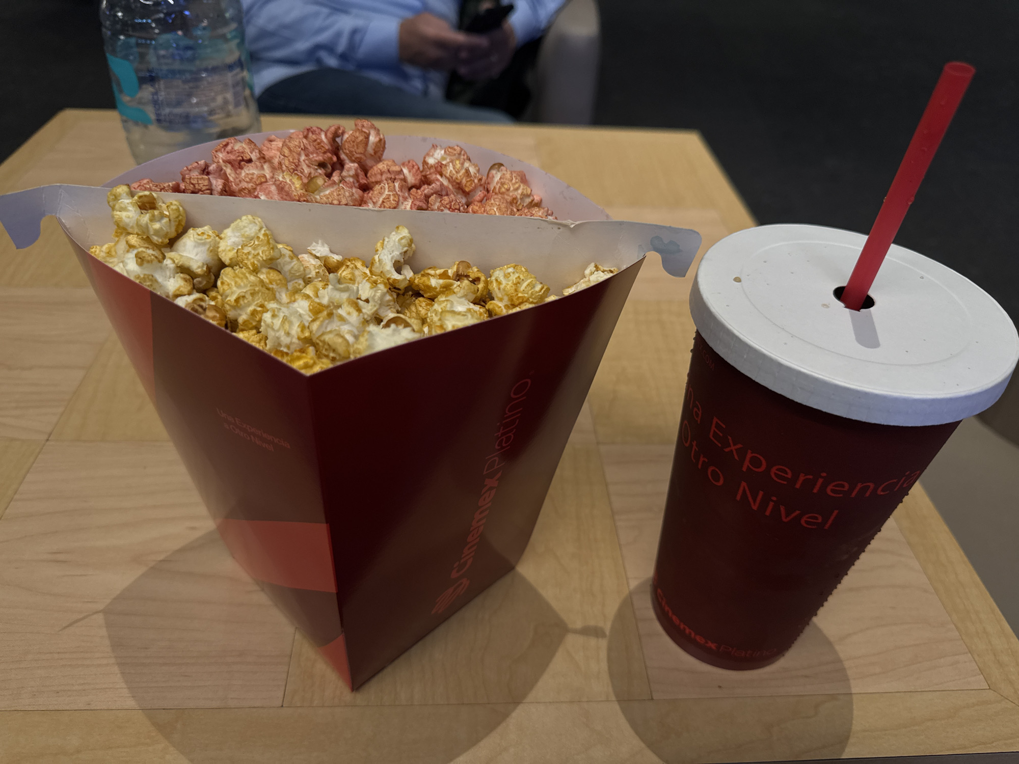 The image captures a cinematic experience in Mexico City, Mexico, featuring a box of Cinepolis Platinum popcorn with two flavors: caramel and strawberry. A Cinepolis Platinum soda with a red straw sits alongside the popcorn. In the background, a person is seated, holding a mobile phone. A water bottle is also visible. The scene is set on a light brown table, presumably inside a Cinepolis Platinum theater.