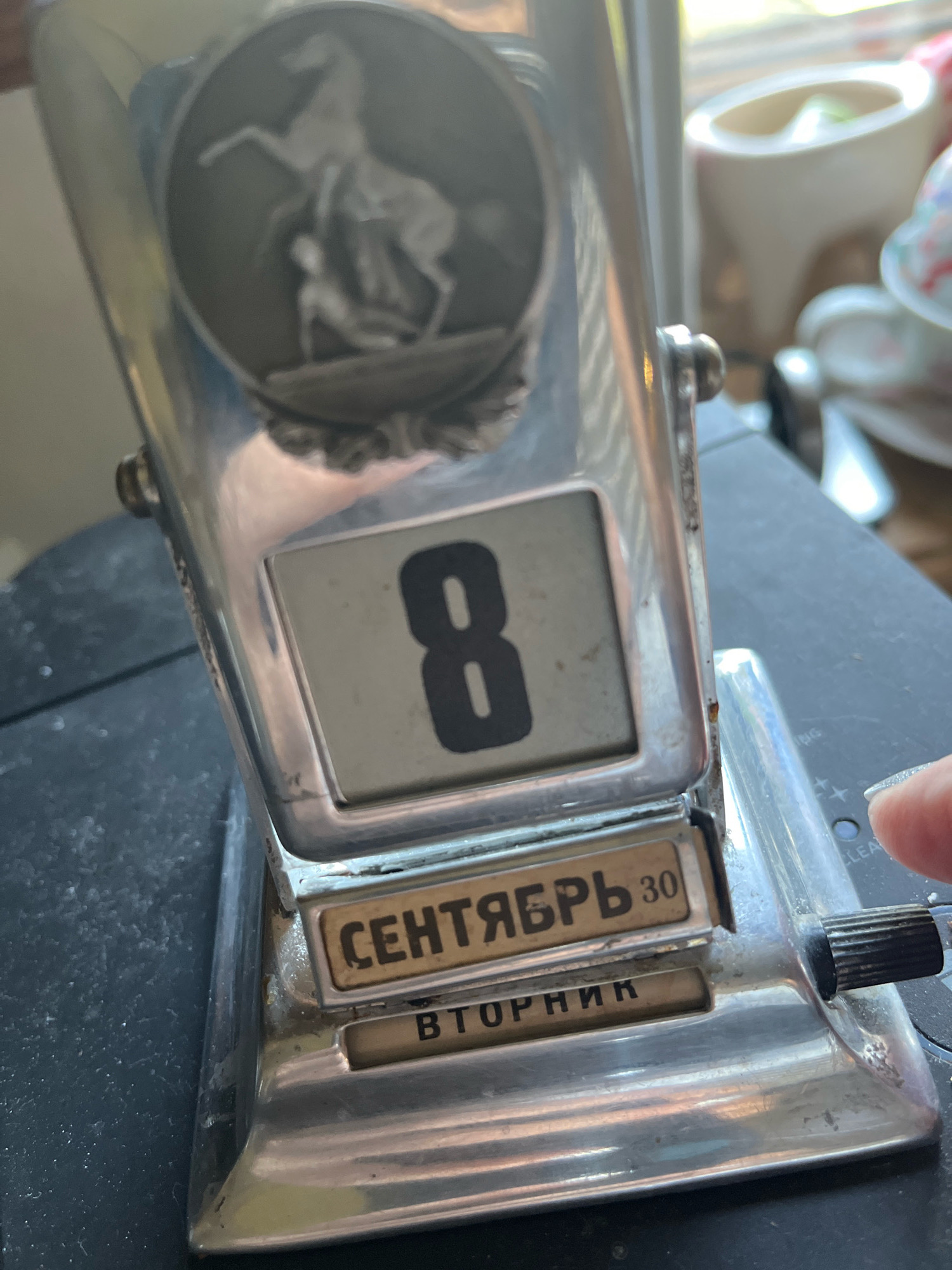 A silver, retro perpetual calendar displays "8" on its digital face. Below, Cyrillic text reads "СЕНТЯБРЬ 30" and "ВТОРНИК," indicating September 30th and Tuesday. The calendar is adorned with an oval medallion featuring a relief of a horse and rider. A hand with manicured fingernails is visible on the right, touching a black dial on the calendar. The calendar rests on a dark, textured surface, possibly a table, with a blurred background showing household items, including a white ceramic pitcher and cups, suggesting an indoor setting in a domestic environment. The lighting is natural, coming from a window to the right.