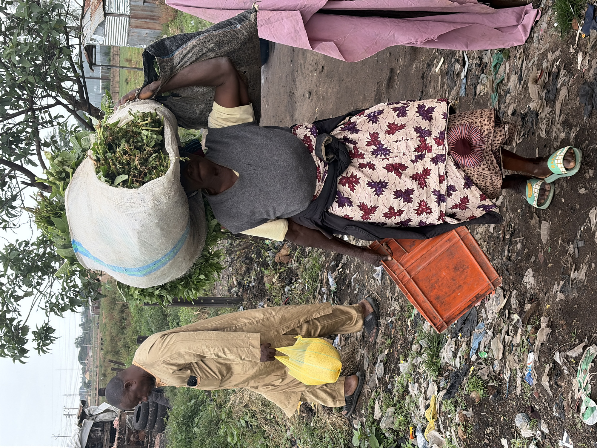 In Bukuru, Nigeria, a woman carries a large sack of green foliage on her head, supported by a second sack over her shoulder. She wears a grey vest over a yellow shirt, a patterned skirt, and light blue sandals. In her right hand, she holds an orange crate. To her left, a man in a tan robe walks with a yellow plastic bag containing a granular substance. The ground is littered with trash, including plastic bags and discarded tires, indicating a possibly impoverished or informal settlement. Buildings with corrugated metal roofs are visible in the background.