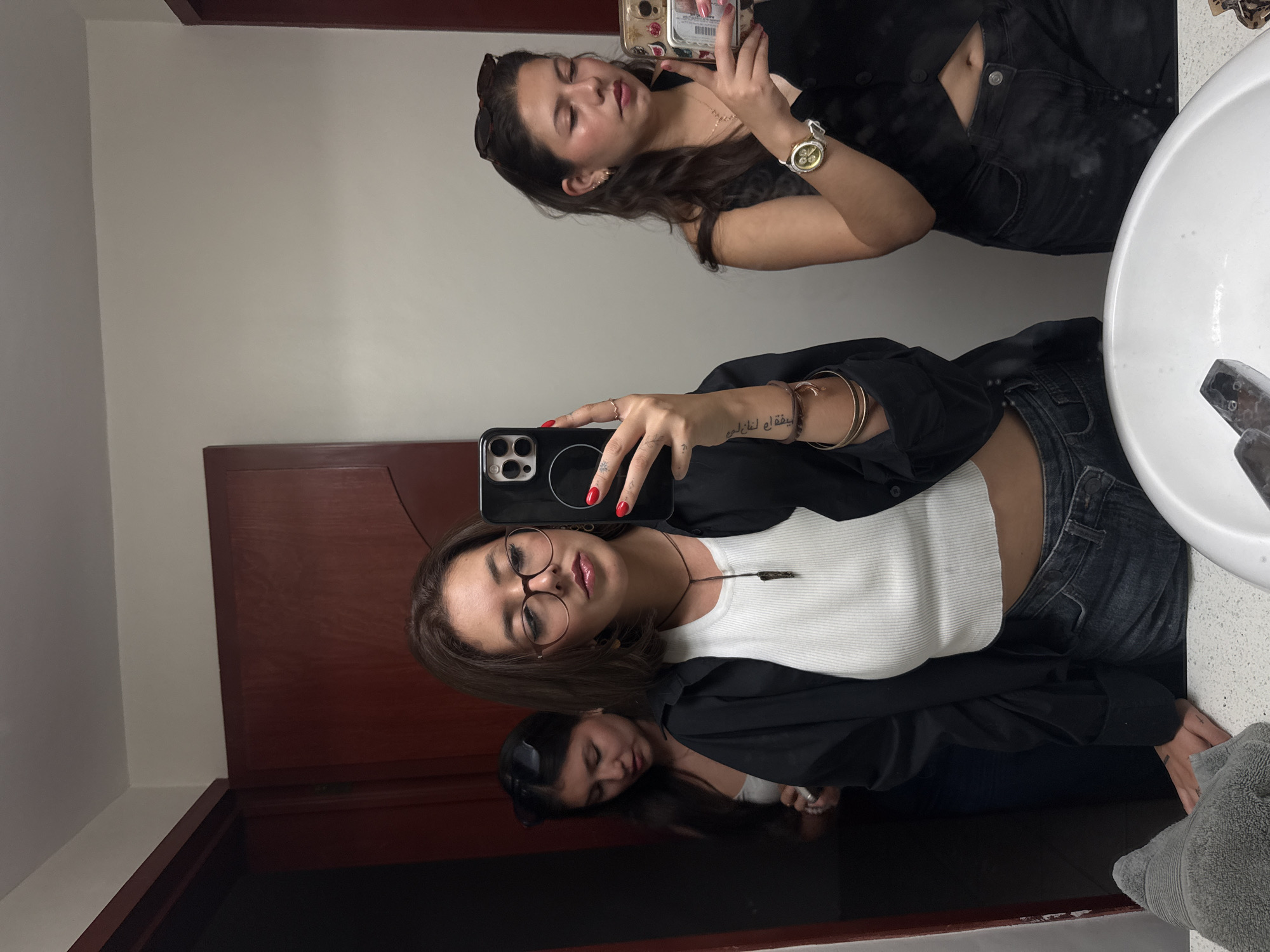 A female takes a selfie in a bathroom mirror. She wears glasses, a white top, and a black jacket. Her reflection shows another female taking a photo, and a third female visible in the mirror. A sink is to the right side of the frame. The photo appears to be taken inside a bathroom in Heroica Puebla de Zaragoza, Mexico.