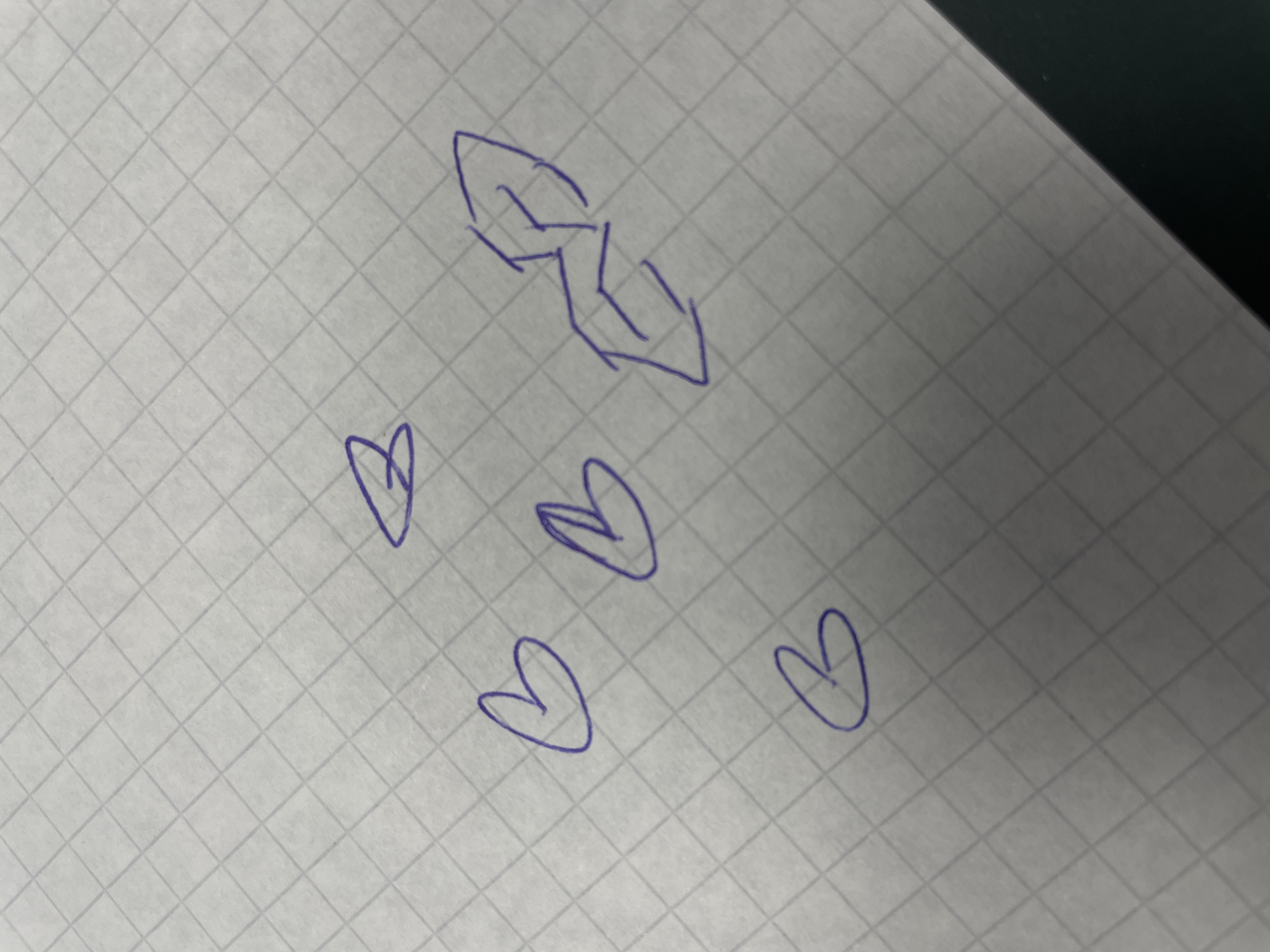 A close-up, slightly angled shot shows a piece of white graph paper with a faint grid pattern. The paper is on a dark surface, possibly a desk. Several doodles are drawn in purple ink. In the upper right quadrant, a stylized drawing resembles a winged creature or an abstract shape made of sharp angles and lines, possibly an insignia or a logo. To the left of this, there are three heart shapes. One is in the upper center, one is to its left and slightly lower, and the third is in the lower left quadrant. There is another heart shape slightly above and to the left of the stylized drawing. The doodles are roughly drawn and do not appear to be related to any specific landmarks or activities in Morsbach, Germany.