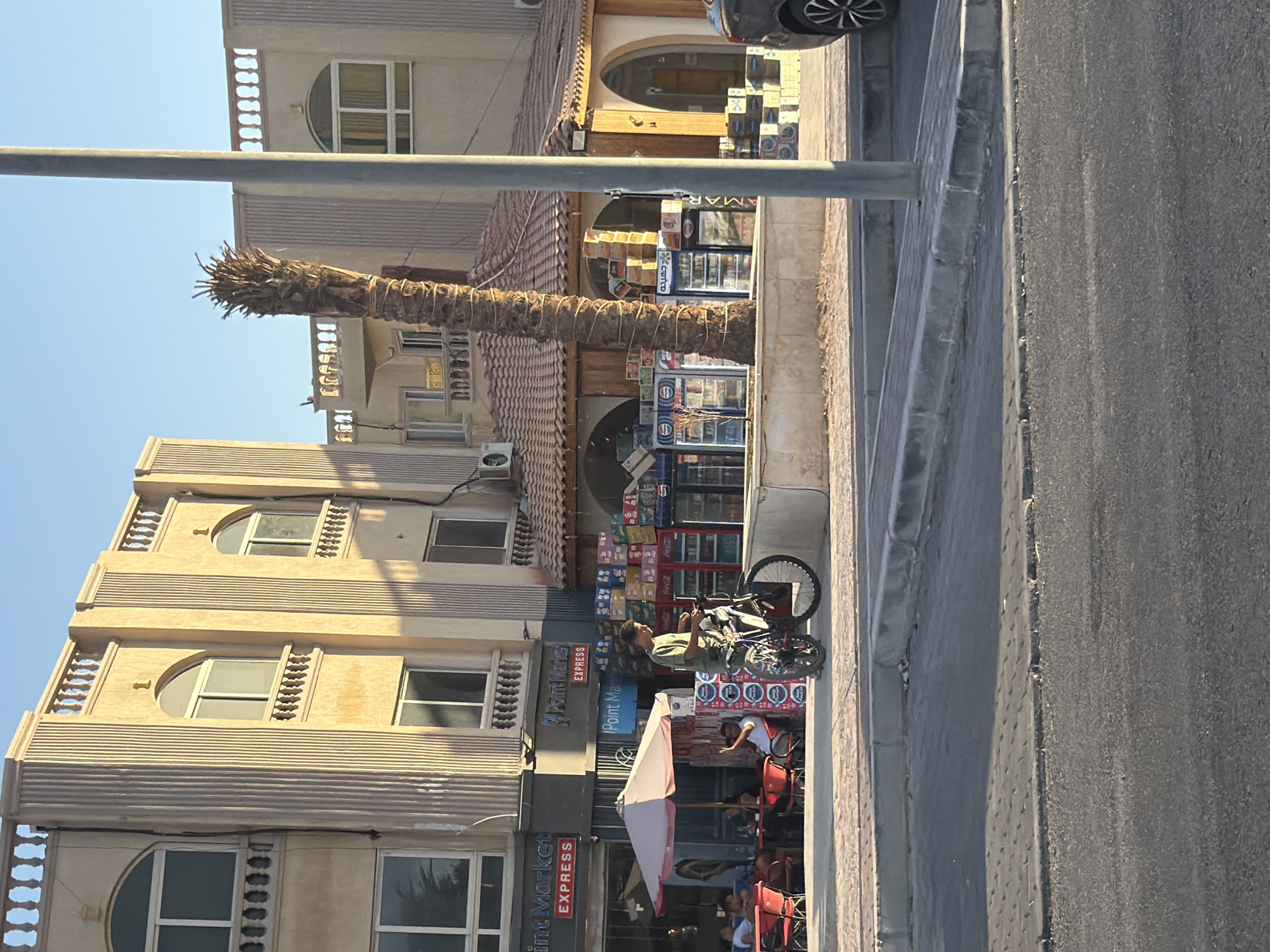 The image depicts a street scene in Sharm El Sheikh, Egypt. The foreground features an asphalt road and a paved sidewalk. A palm tree trunk, denuded of its fronds, stands prominently. To the left, a multi-story building with light-colored facade and decorative balconies lines the street. Windows are visible on various floors, some with rounded tops.
In the mid-ground, several shops are open. One shop has refrigerators stocked with beverages, and a display of boxed items above. Another shop sign reads "Point Market EXPRESS" in English. A person is sitting on a bicycle, looking towards the shops. Further down, an umbrella shades an outdoor seating area with chairs and tables, where a few people are seated. The sky above is clear and blue, suggesting good weather.
The time of day appears to be late afternoon or early evening, indicated by the angle of the sunlight casting shadows. Visible text includes shop names like "Point Market EXPRESS" and brand names such as "جهينه" (Juhayna) and "AMAR". The overall impression is that of a busy commercial street in a resort town.