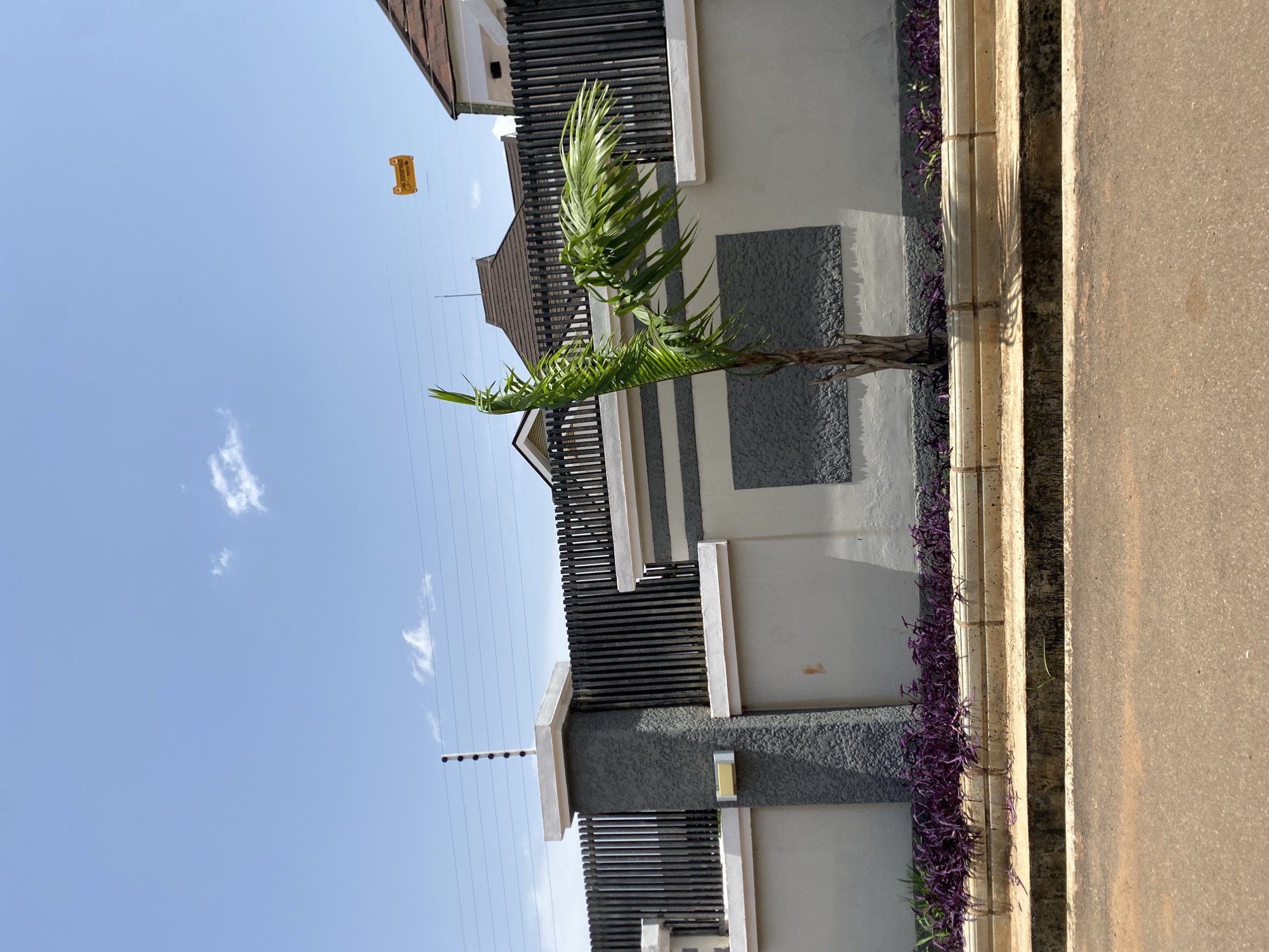 The image depicts a section of a modern fence with an electric deterrent system on top. The fence is constructed with grey concrete pillars and a dark vertical slat design, interspersed with light grey panels. Below the fence, a row of vibrant purple plants lines the base. A small palm tree stands to the right of the central pillar. The sky above is clear blue with a few wispy clouds. A yellow warning sign is visible on a wire strung across the sky. The foreground shows a paved road and a concrete drainage channel. This scene is located in Gura Top, Nigeria.