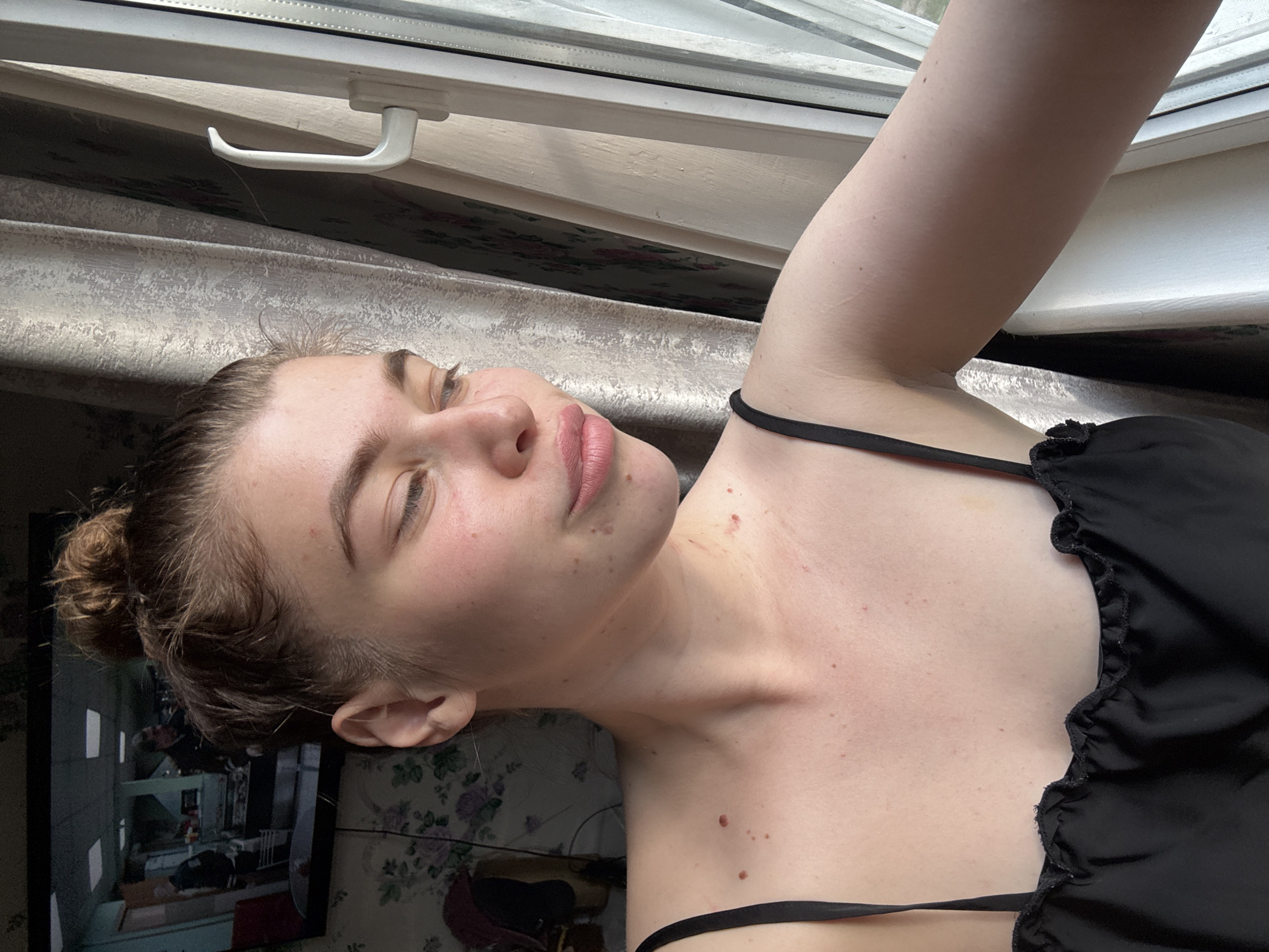 A fair-skinned woman with brown hair pulled into a messy bun is lying on a brown, patterned surface with her head near a window. She wears a black top with a spaghetti strap. The window, with a white frame and handle, is partially visible in the upper portion of the frame, while a floral wallpaper can be seen in the lower left corner. The image is tilted, suggesting it was taken at an angle. The location is not confirmed to be Мещёры, Russia.